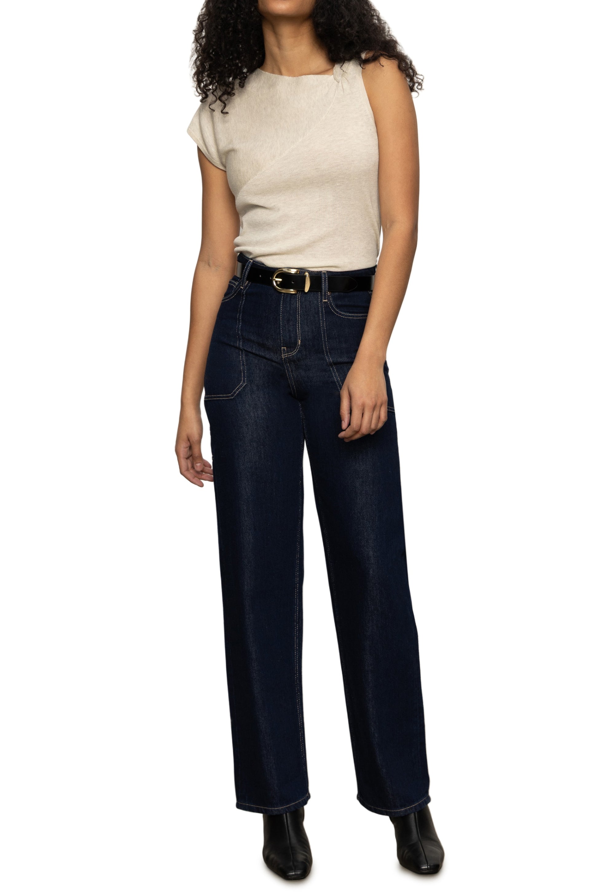 A person in a beige Sanctuary Asymm Shoulder Twist Top, dark blue high-waisted jeans with a black belt, and black ankle boots stands against a white background. Their face is not visible.