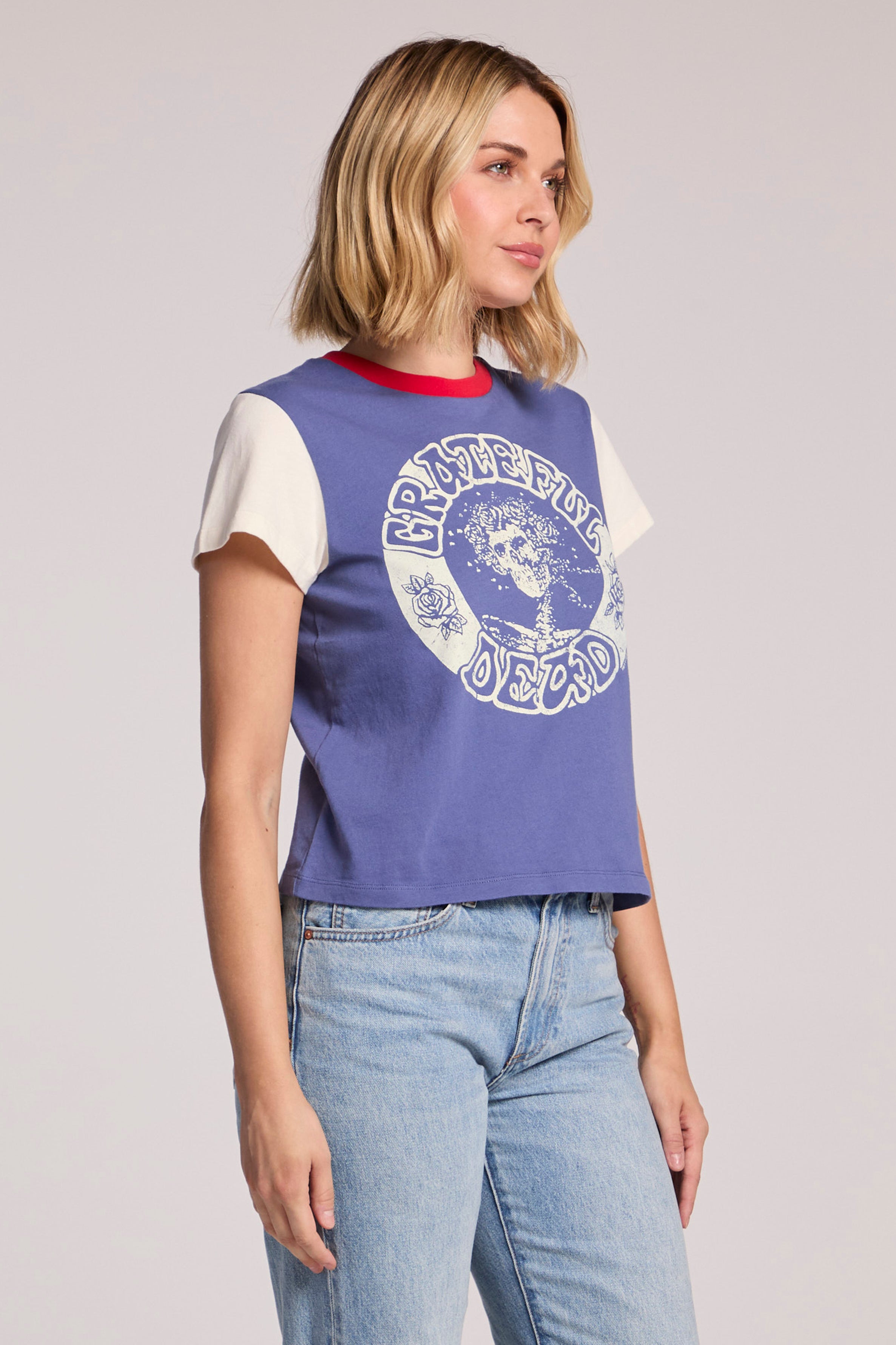 A blonde woman in the Chaser Grateful Dead - Classic Bertha tee with cream sleeves and a Bertha skull graphic, paired with blue jeans, stands against a plain background, looking to the side.