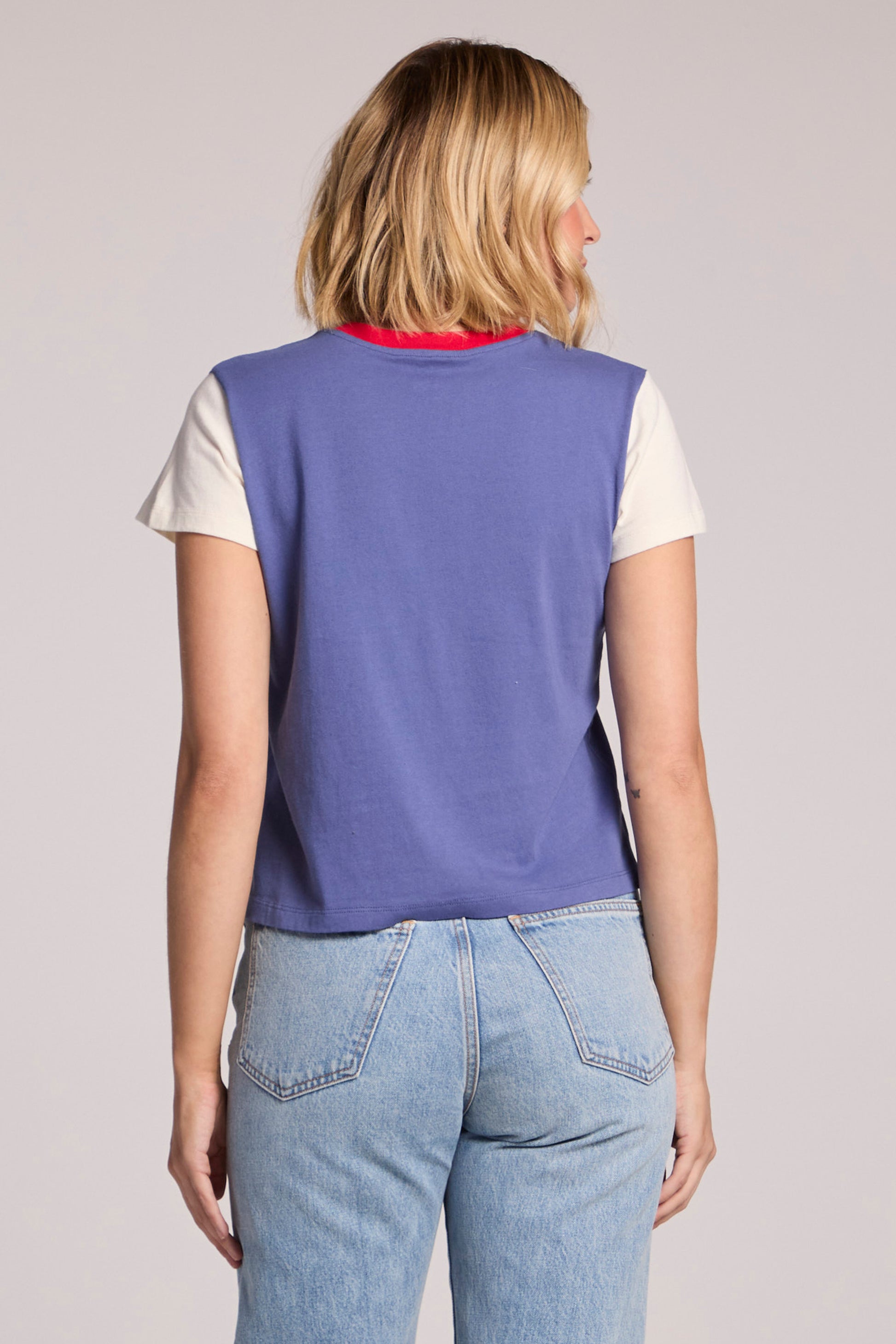 A woman with shoulder-length blonde hair, in light blue jeans and a Chaser Grateful Dead - Classic Bertha tee, featuring purple, white, and a red neckline, stands with her back to the camera against a plain background.