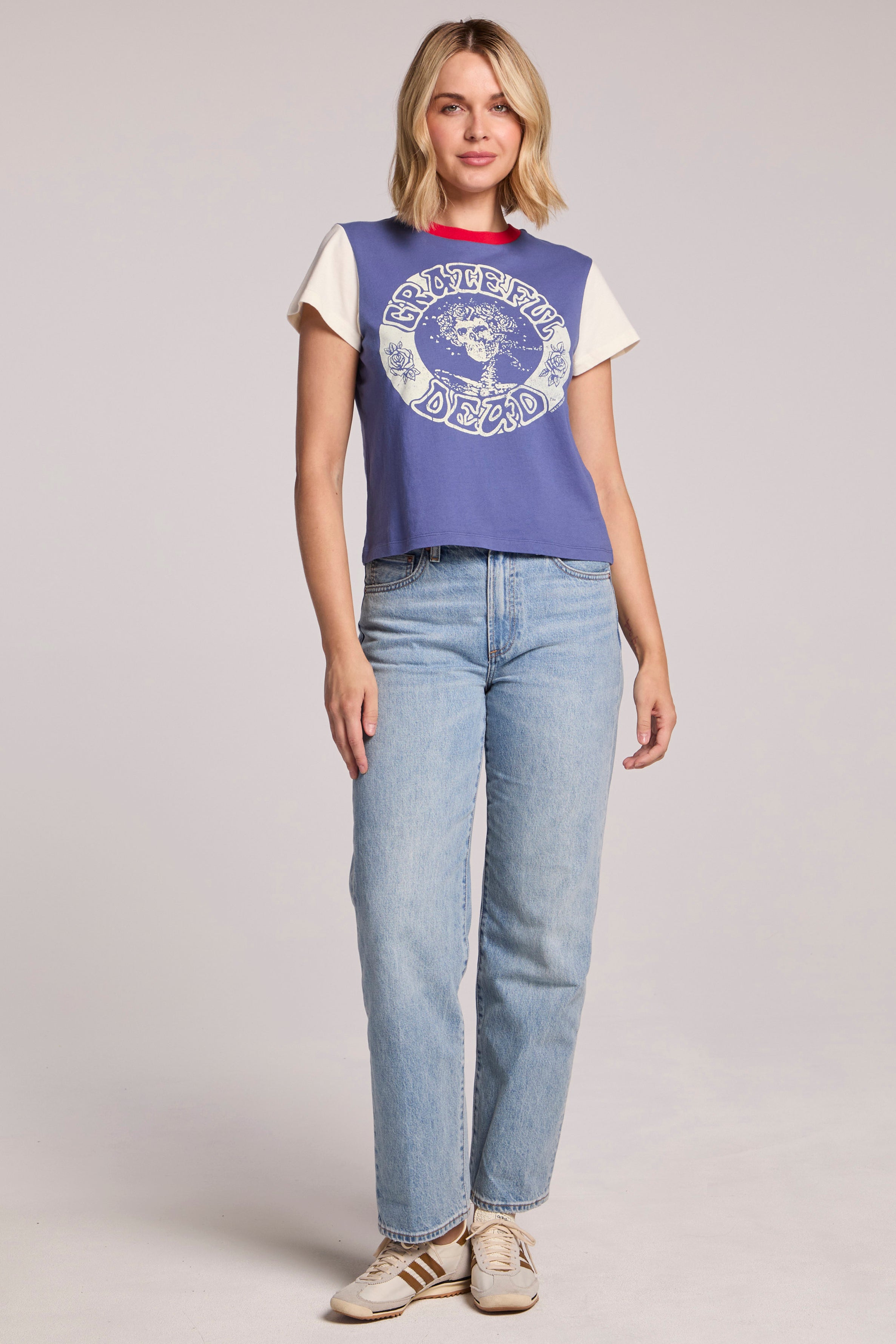 A blonde woman faces forward, wearing the Chaser Grateful Dead - Classic Bertha tee in blue and white, light blue jeans, and beige Adidas sneakers against a plain light background.