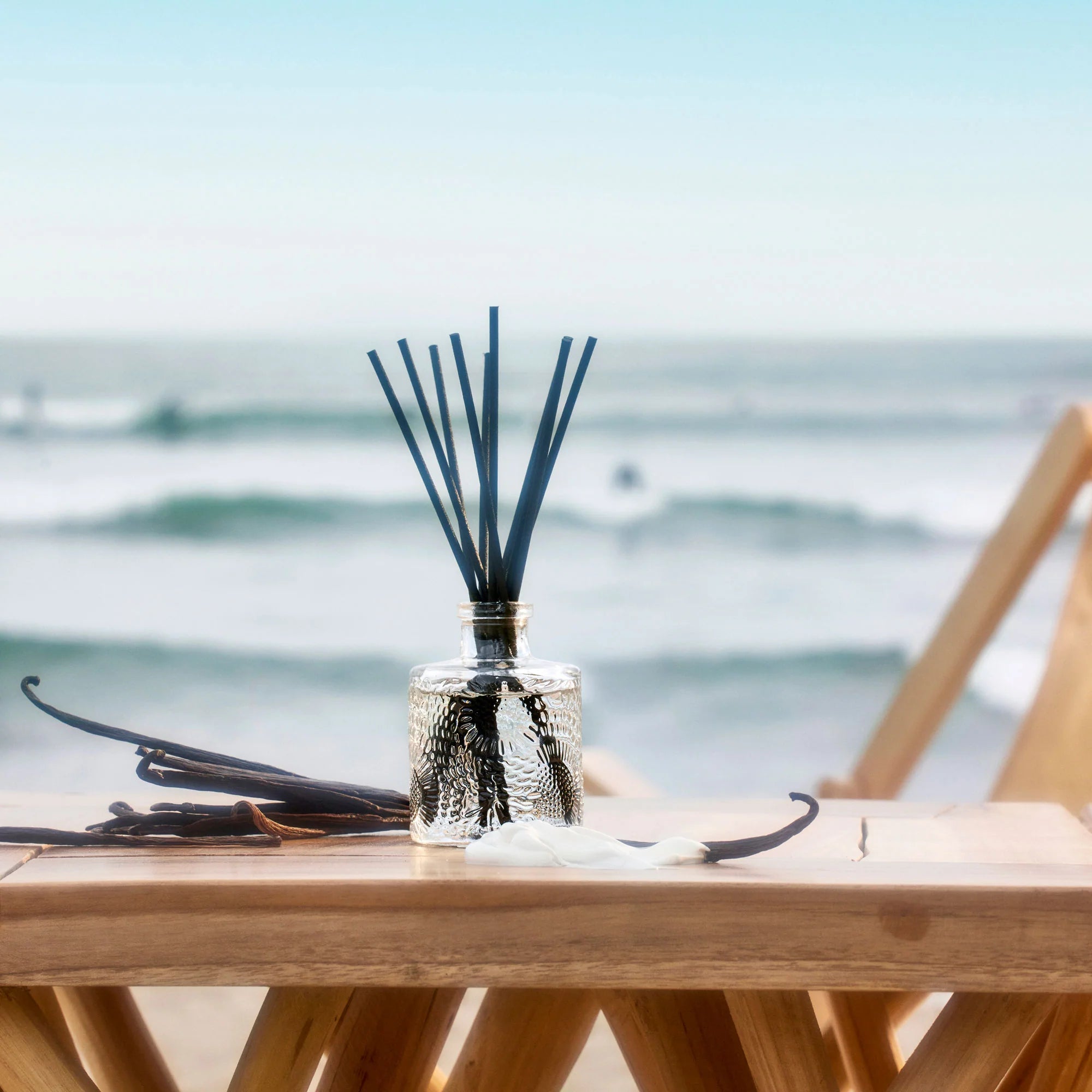 The Voluspa California Summers Large Diffuser (3.4 fl oz) offers a vegan scent experience, featuring vanilla notes and a white flower, set on wood with ocean waves in the background, lasting throughout the diffuser’s life.