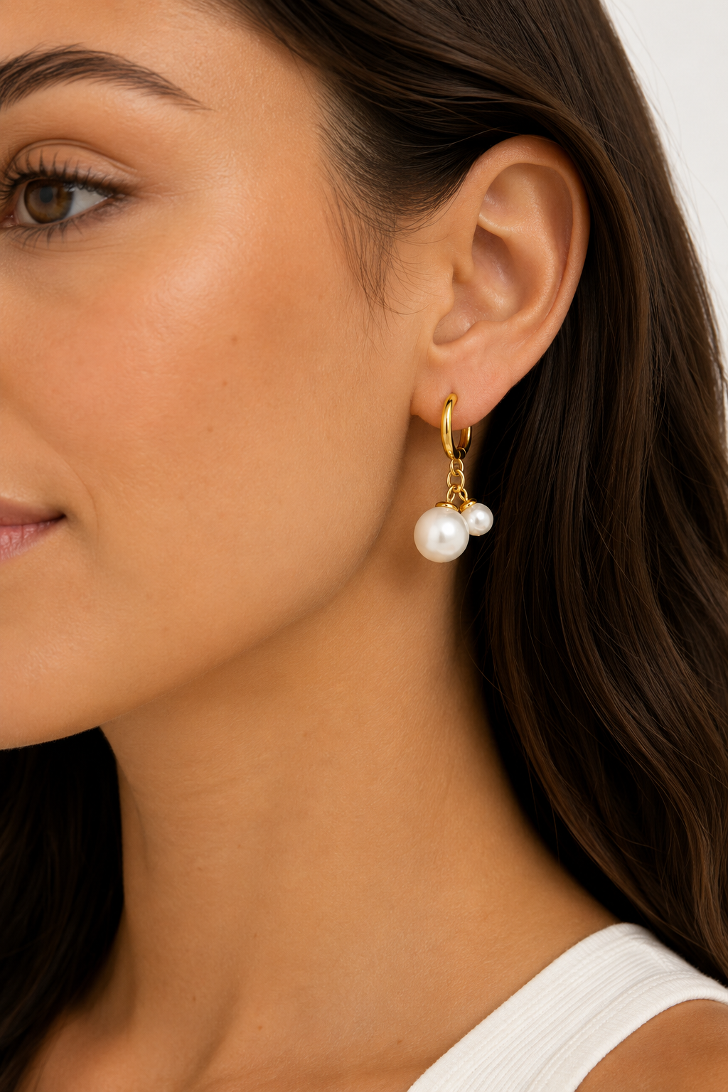 Close-up of a woman wearing Secret Box’s Water Resistant Pearl Huggie Hoop on her left ear, featuring gold hoops with one large and one small white pearl. She has smooth skin, long brown hair, and a white top.