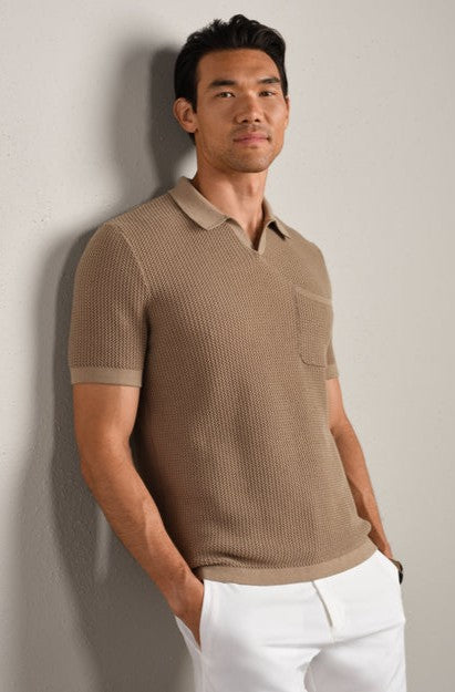 A man with short black hair and a neutral expression stands against a light wall, wearing the South Moon Under Coastal Johnny Collar Polo in Tan Multi with white pants. The polo features classic fit, ribbed trim, knit stitching, and a chest pocket.