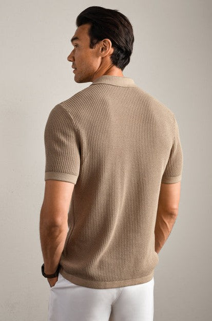 A man with dark hair, seen from behind, wears the Coastal Johnny Collar Polo by South Moon Under—it's short-sleeved, beige, textured with ribbed trim—paired with white pants against a plain, light background.