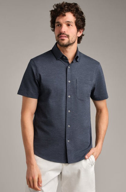 A man with wavy brown hair and a beard wears the South Moon Under Commuter Seersucker Short Sleeve Shirt in navy, paired with white pants. He stands against a light gray background, looking slightly to the side.