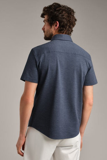 A man with short, curly brown hair is seen from behind wearing South Moon Under’s Commuter Seersucker Short Sleeve Shirt in navy blue and white pants, standing against a plain gray background.