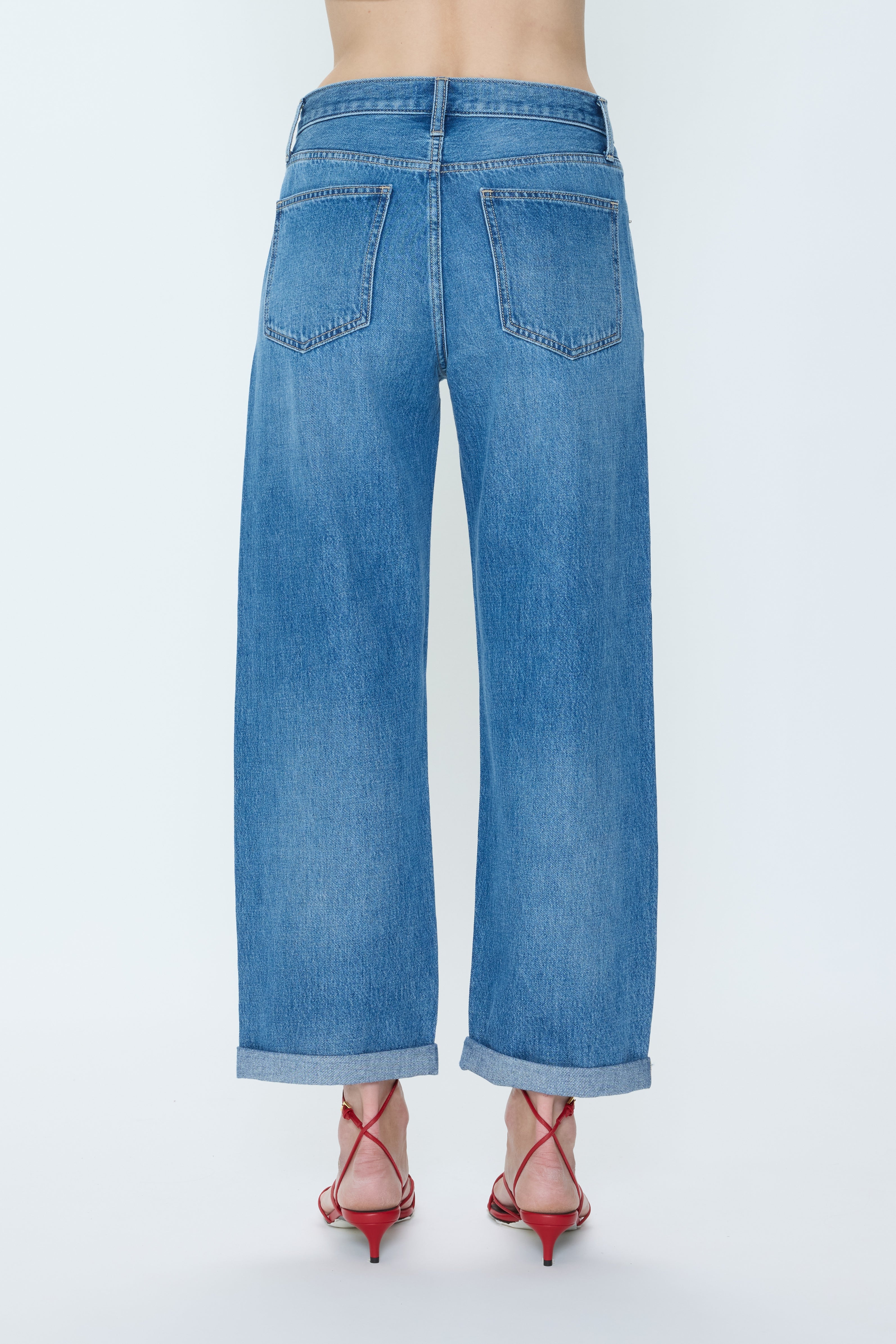 A person stands with their back to the camera, wearing Pistola Donny Rolled Hem Bf high-waisted wide-leg blue jeans with rolled cuffs and red strappy heels against a plain light background.
