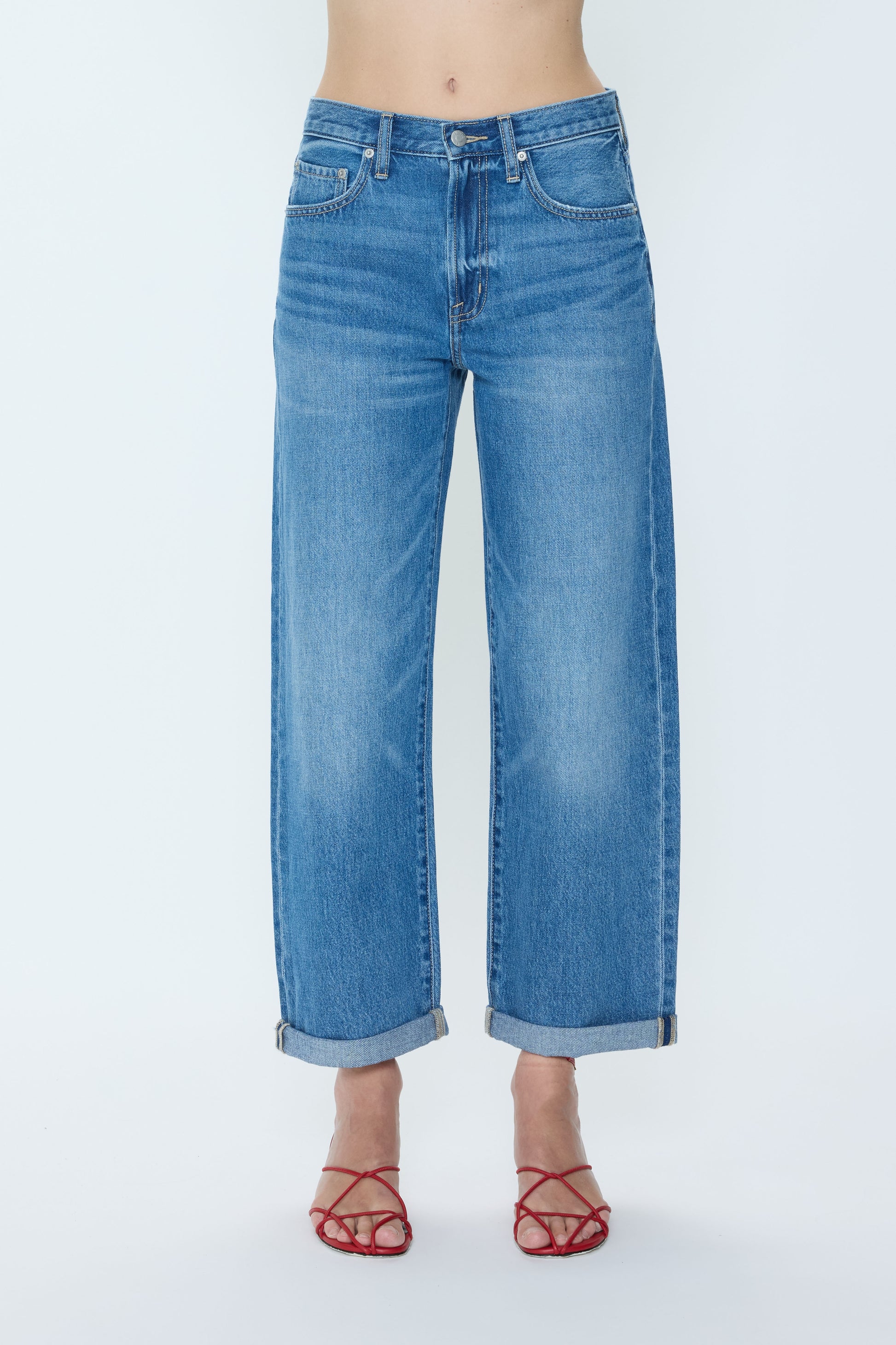 A person wearing Pistola Donny Rolled Hem Bf jeans in HEARTFELT, styled with rolled cuffs and paired with red strappy sandals, stands against a plain white background.