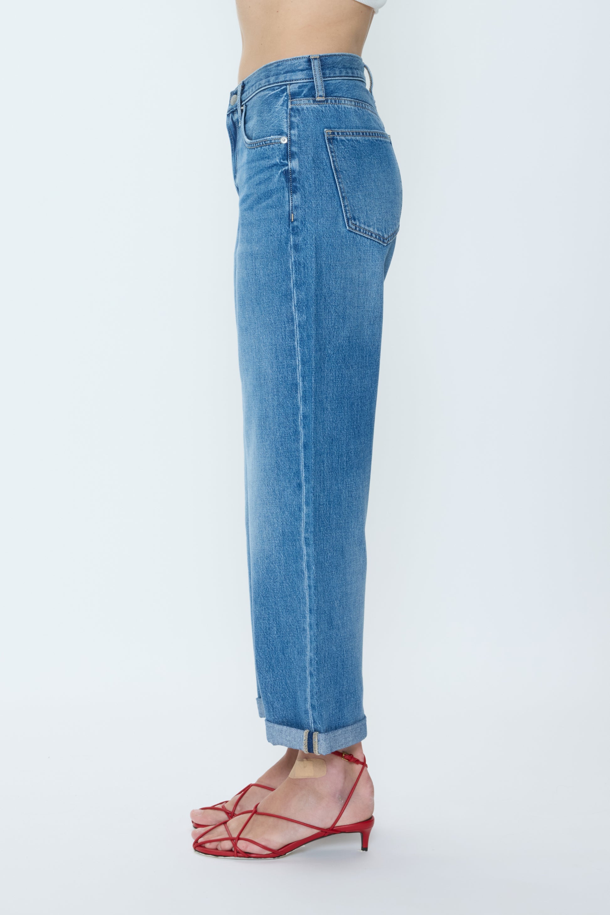A person stands sideways against a plain light background, wearing Pistola Donny Rolled Hem Bf light blue relaxed boyfriend-fit mid-rise 100% cotton jeans with red strappy heeled sandals; upper body is partially visible.