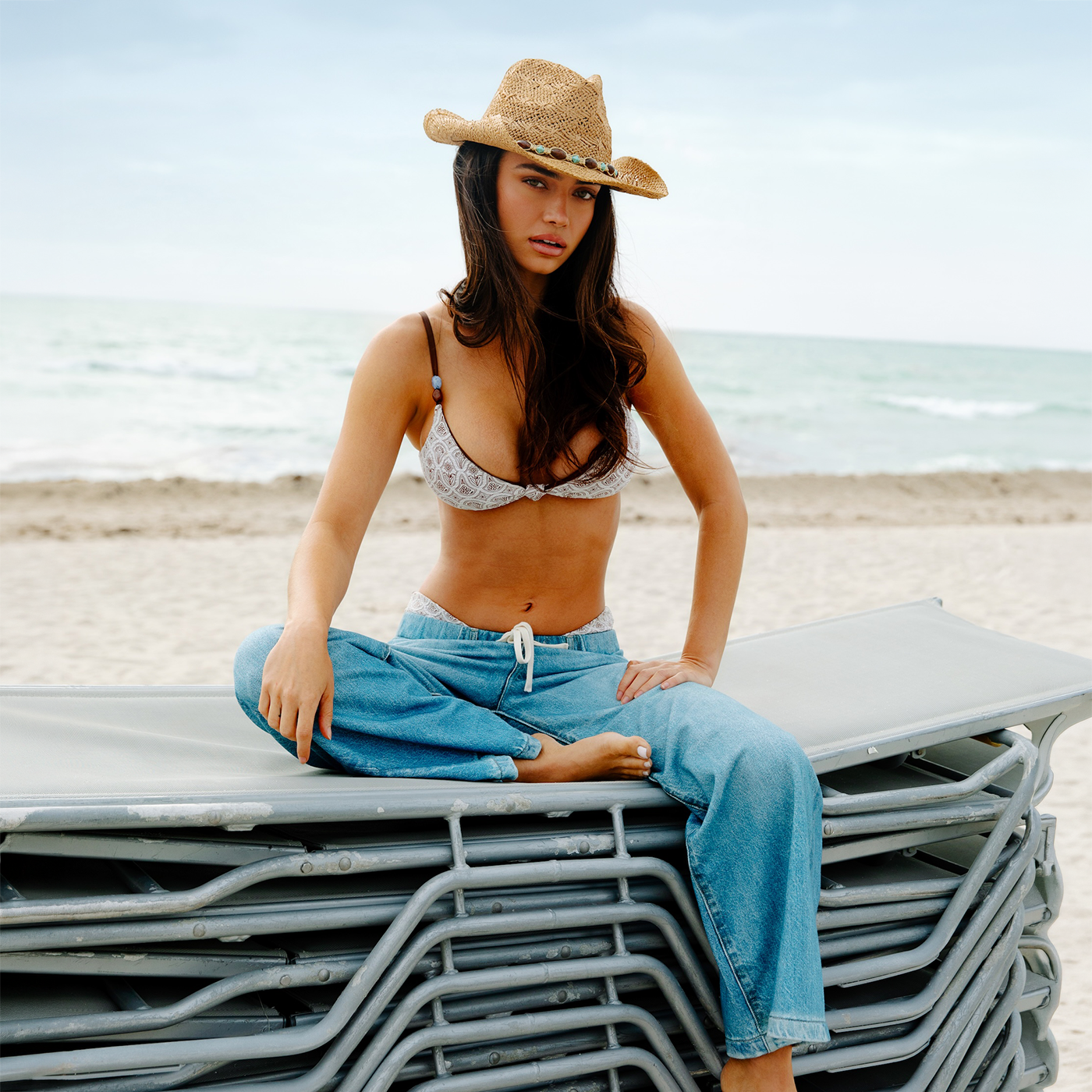 A woman in a straw hat and Dippin' Daisy’s Samba & Smocked Flora Zen Top sits cross-legged on stacked lounge chairs at the beach, with the ocean and sky behind her.
