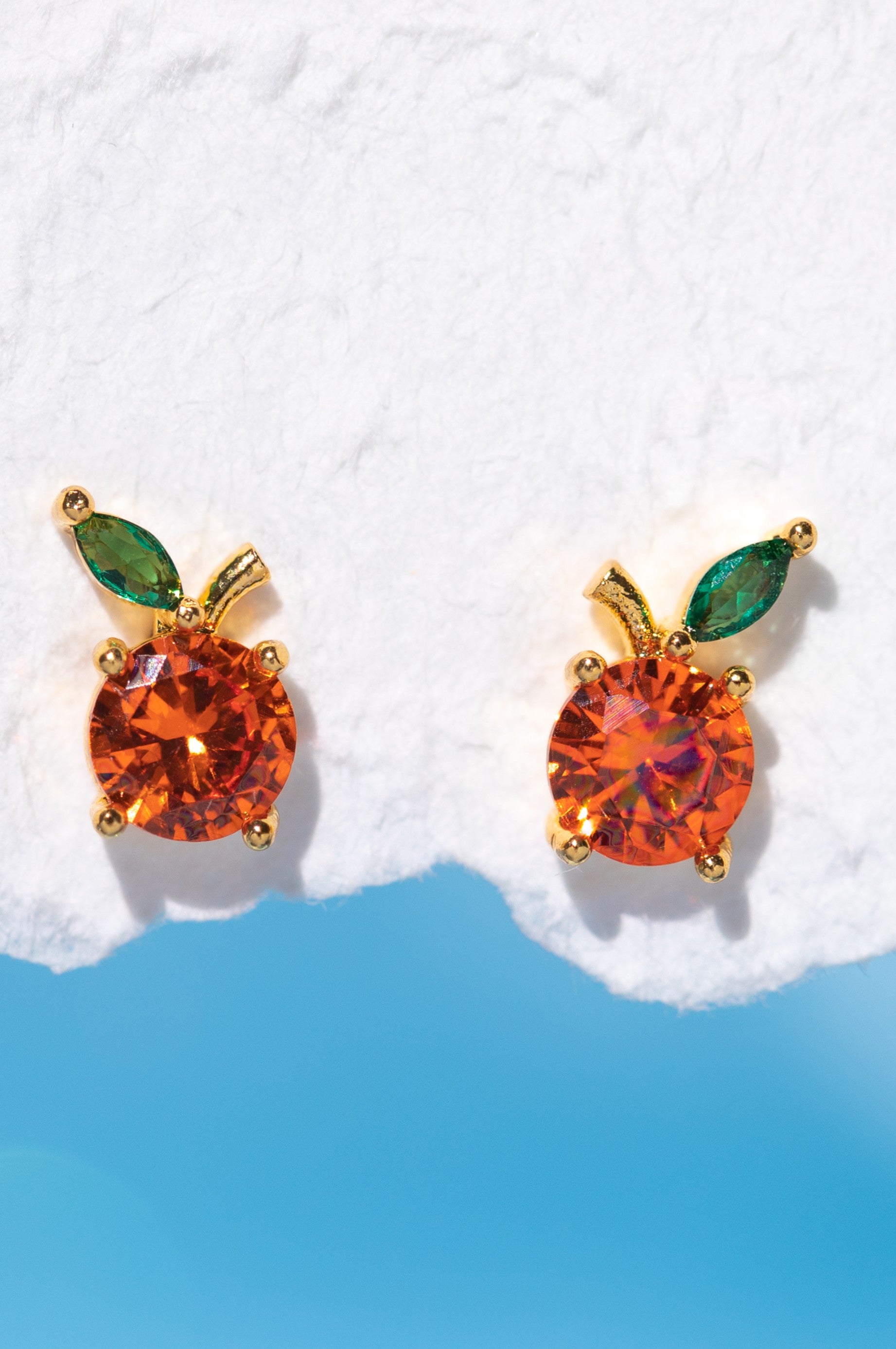 The Girls Crew Orange Juice Stud Earrings, crafted with 18k gold-plated finishes and sterling silver posts, feature orange-colored cubic zirconia as fruit and leaf details, displayed on a white textured surface above a blue background.