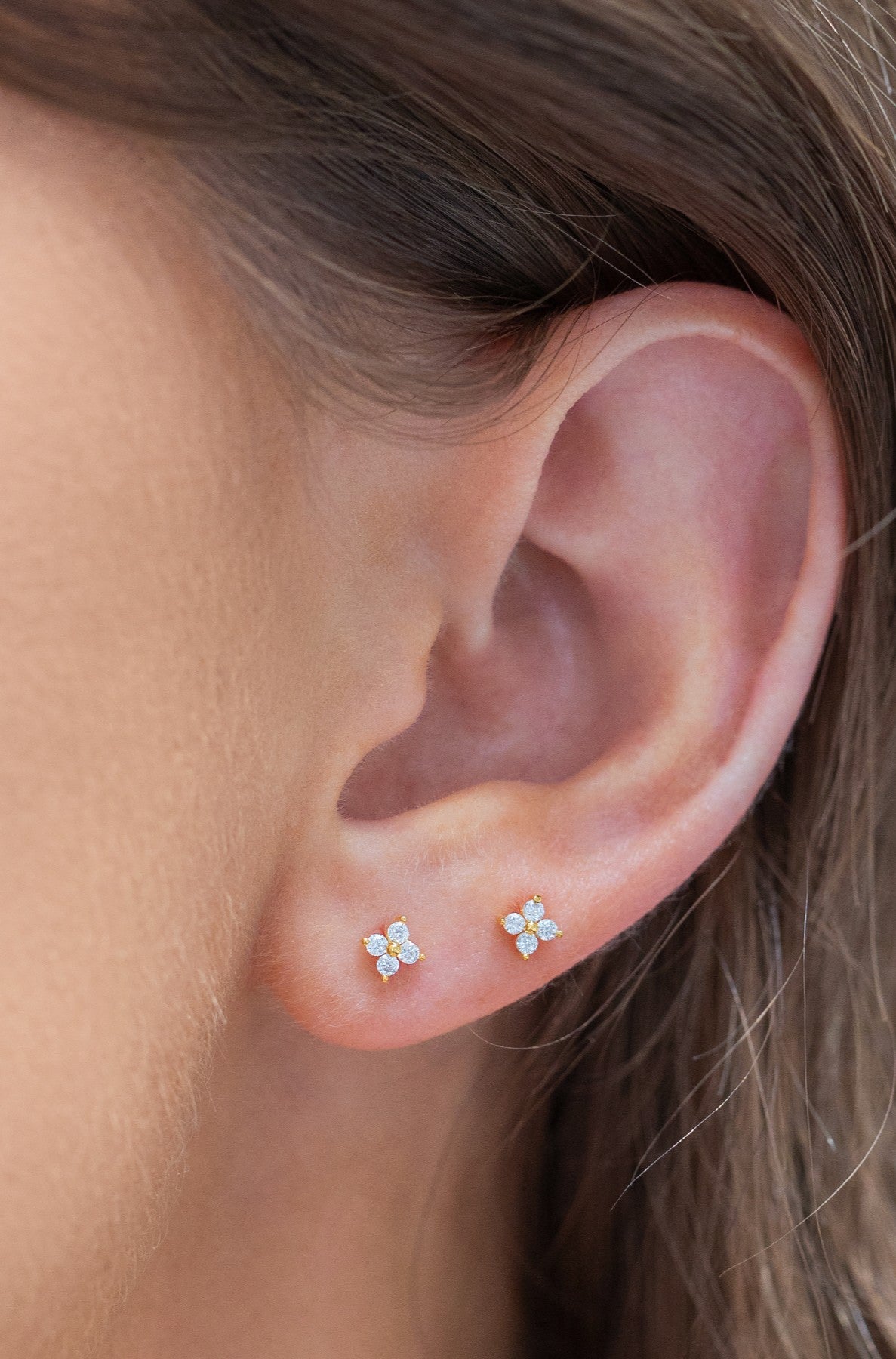 Close-up of a woman's ear wearing Girls Crew Teeny Tiny Clear Cluster Studs: two petite, four-petaled earrings with sparkling cubic zirconia centers, 18k gold plating, and sterling silver posts for comfort.