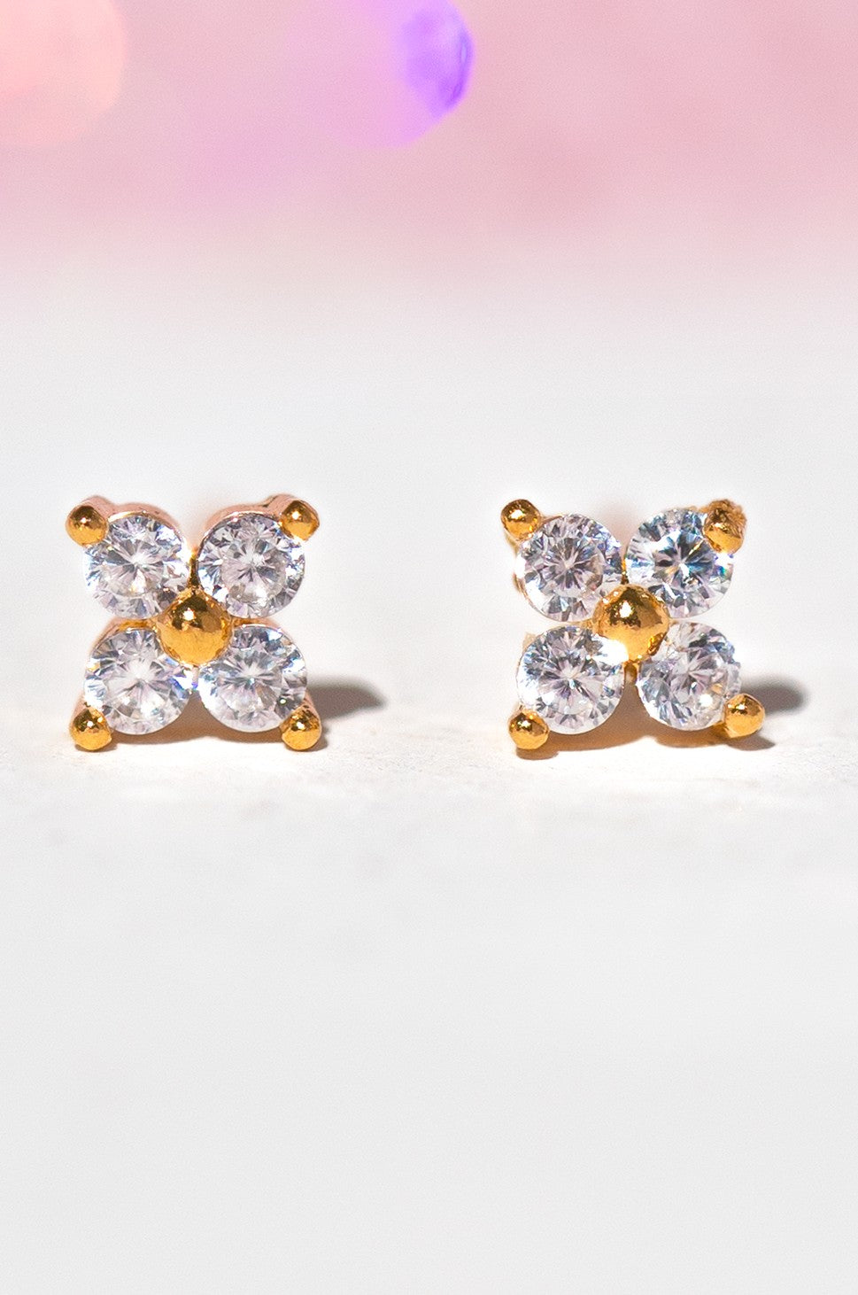 The Girls Crew Teeny Tiny Clear Cluster Studs are gold stud earrings, each with four round clear cubic zirconia stones in a square pattern, shown on a soft pink and white background.
