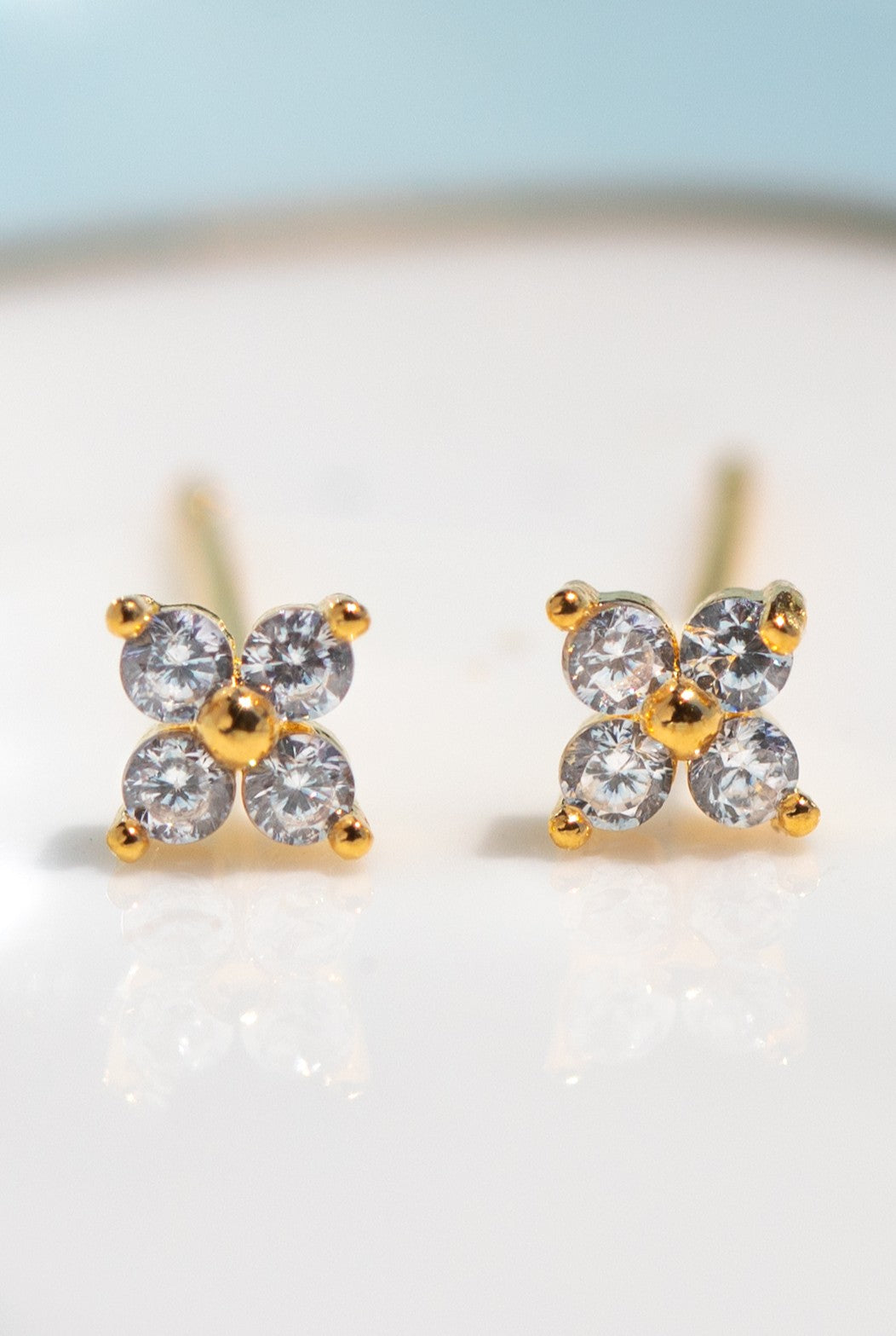 Close-up of Girls Crew Teeny Tiny Clear Cluster Studs: 18k gold plated stud earrings with sterling silver posts, each adorned with four round cubic zirconia in a floral design, shown on a white surface.