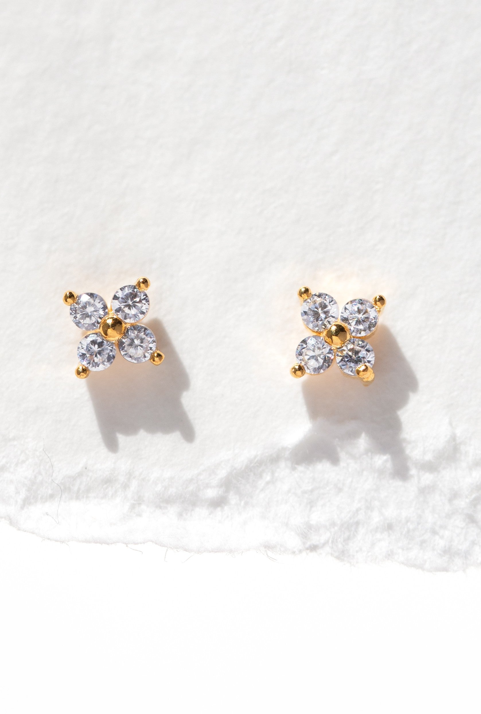 Girls Crew Teeny Tiny Clear Cluster Studs: 18k gold plated flower stud earrings with four clear cubic zirconia petals and sterling silver posts, shown on a textured white background.