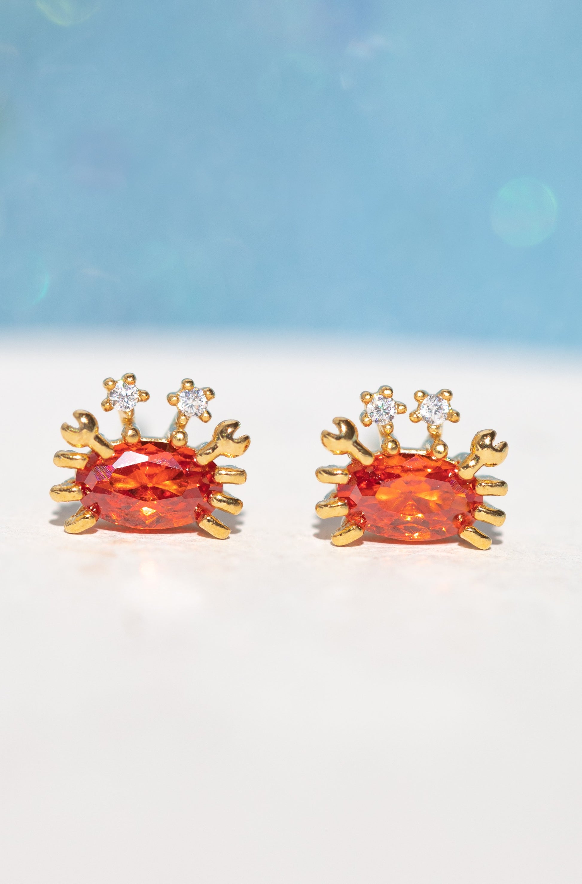 Girls Crew's Cutie Crab Stud Earrings feature two red crab-shaped studs with orange gemstone centers and cubic zirconia eyes, crafted with 18k gold plating and sterling silver posts, shown on a soft blue and white background.