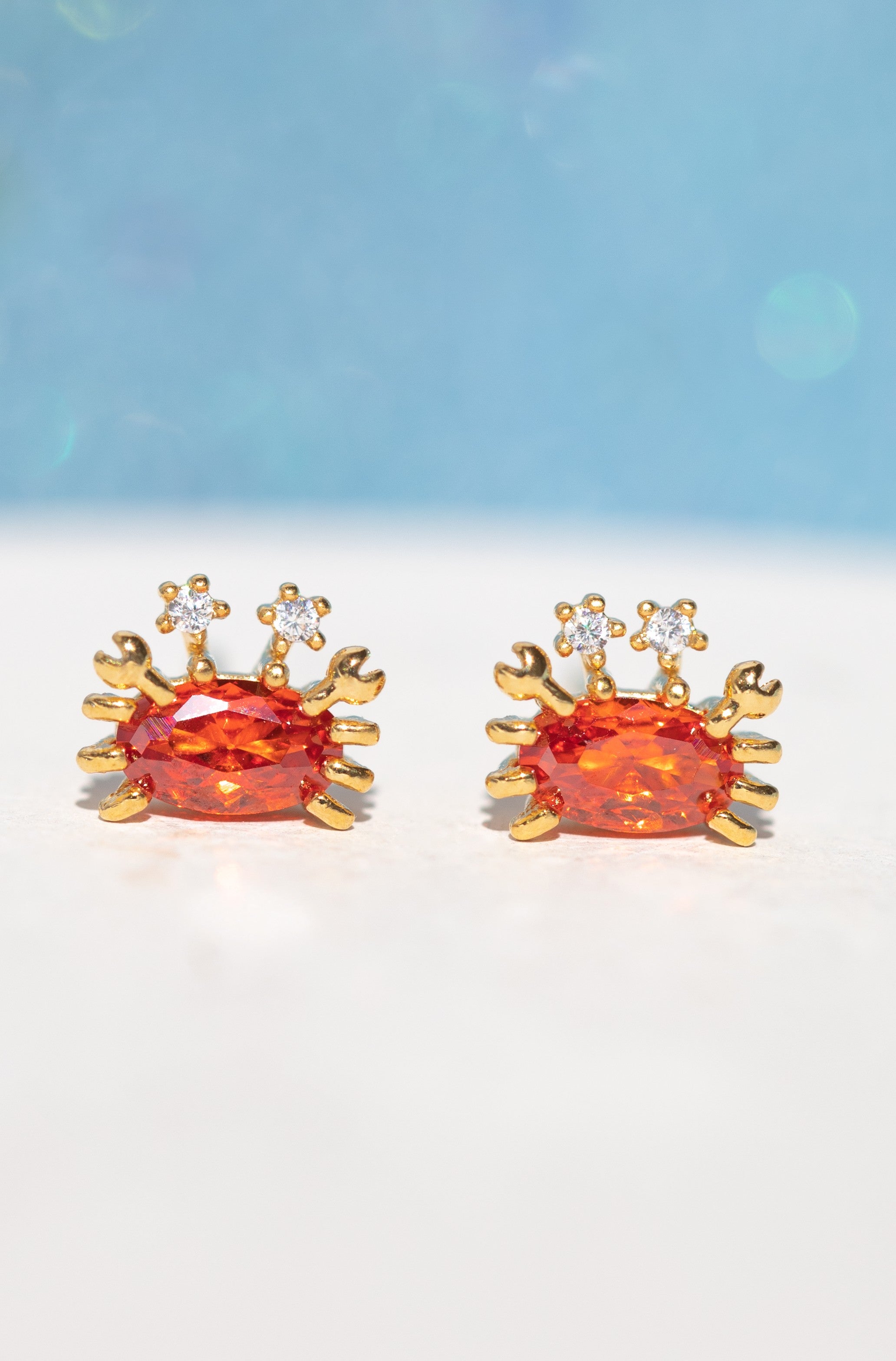 Girls Crew's Cutie Crab Stud Earrings feature two red crab-shaped studs with orange gemstone centers and cubic zirconia eyes, crafted with 18k gold plating and sterling silver posts, shown on a soft blue and white background.