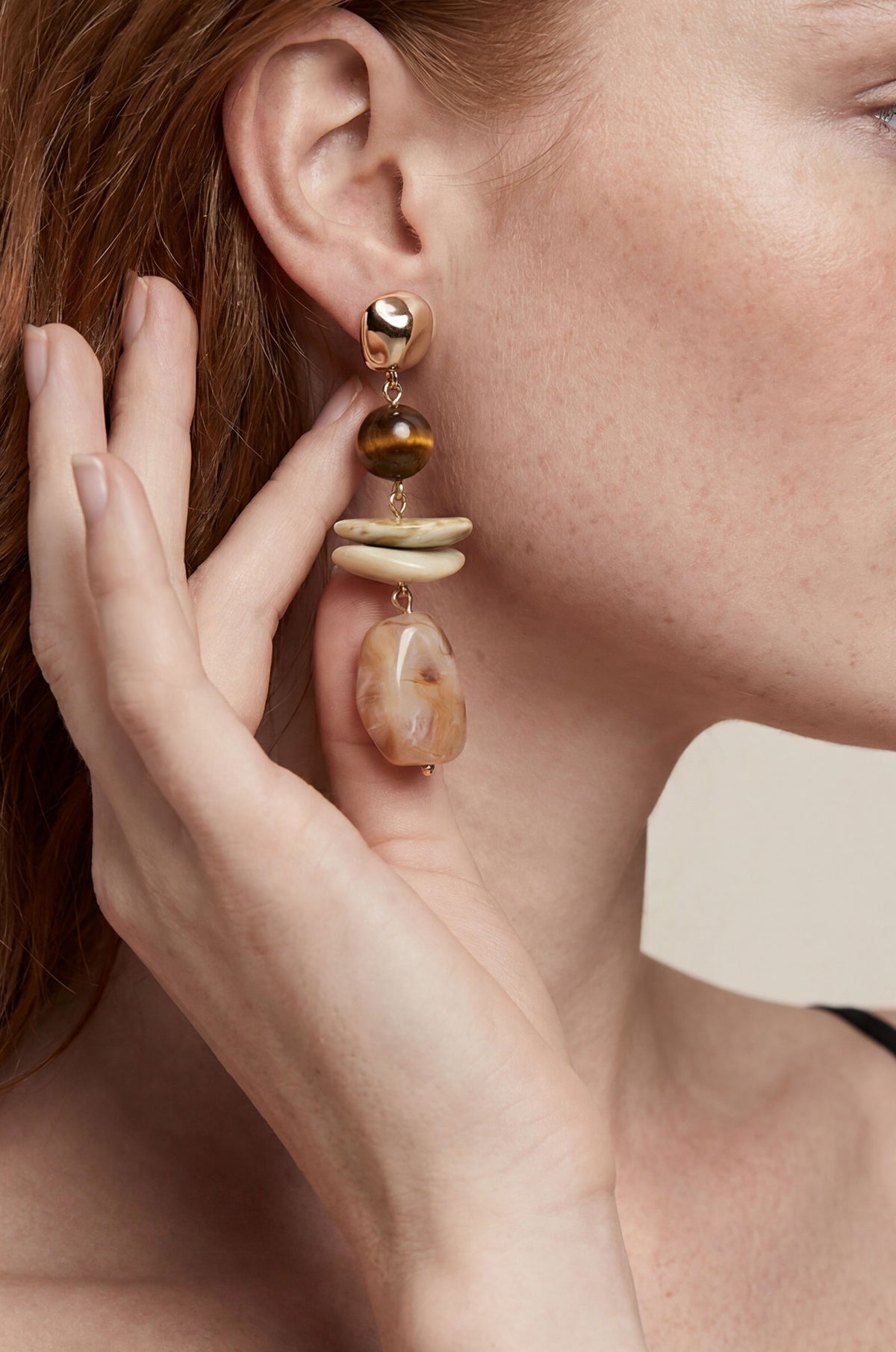 Gold-tone drop earrings with brown stone bead, cream-colored spacer elements, and translucent amber stone pendant worn on ear