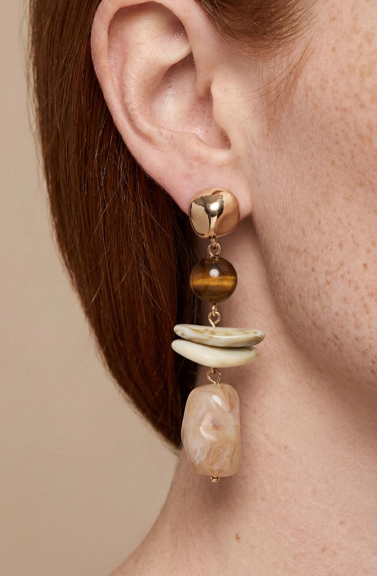 Gold-tone drop earring with tiger's eye bead, cream curved element, and beige stone pendant.