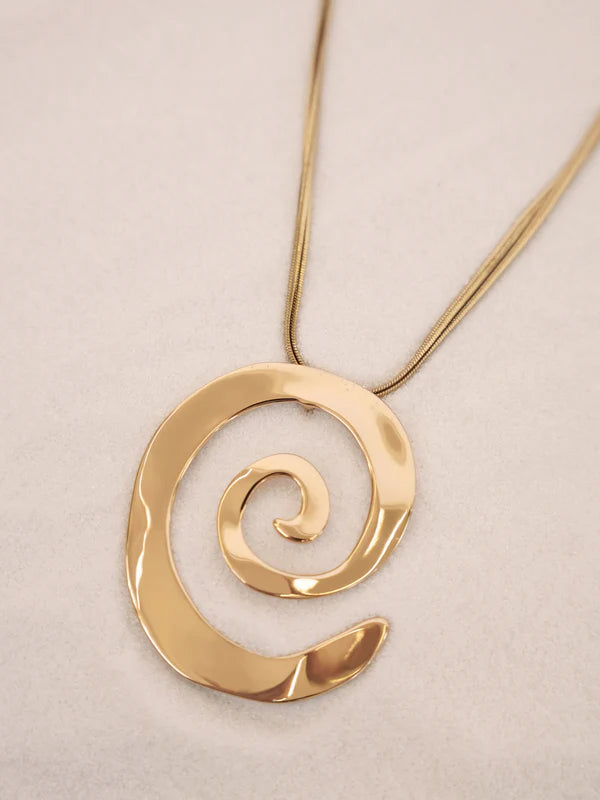 The Ettika Aura Swirl Necklace features a large, shiny spiral-shaped pendant with 18k gold plating, displayed on a light beige surface.