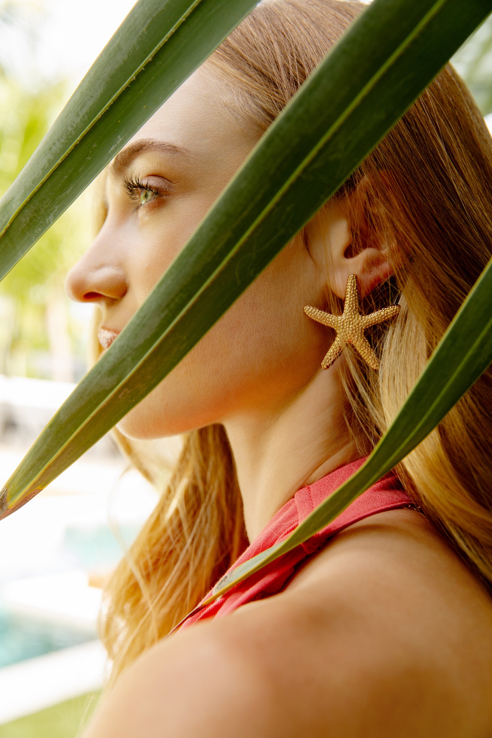 A woman with long blonde hair, in a pink top, wears Ettika Statement Starfish Stud Earrings in gold while partially hidden by large green leaves in natural outdoor light.