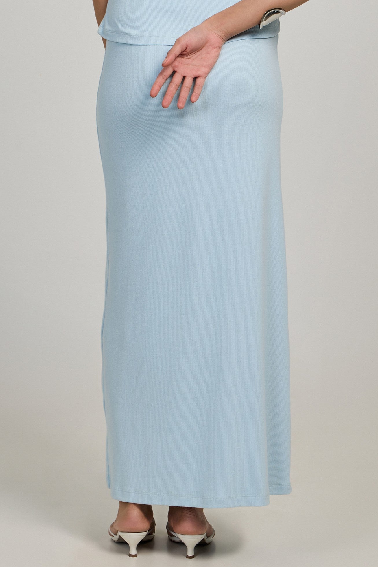 A person in the Summer Away Minato Skirt, a full-length light blue ankle-length style, stands with their back to the camera, wearing white heeled sandals and resting one hand behind them with fingers crossed.
