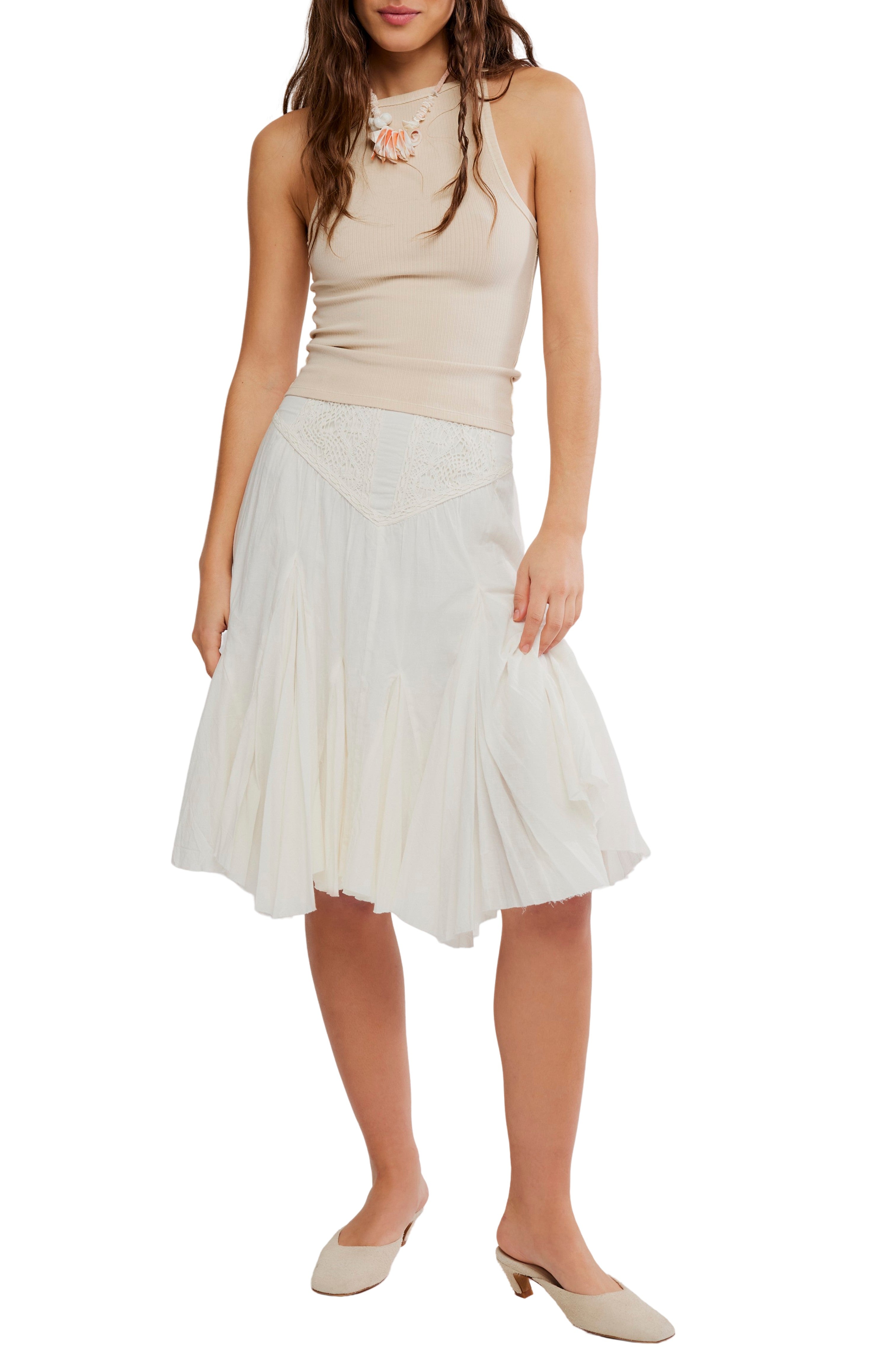 A woman stands against a white background wearing a sleeveless beige ribbed top, beige low-heeled shoes, a necklace, and the Free People Fp 1 Frida Godet Midi Skirt—a white high-rise skirt with lace details.