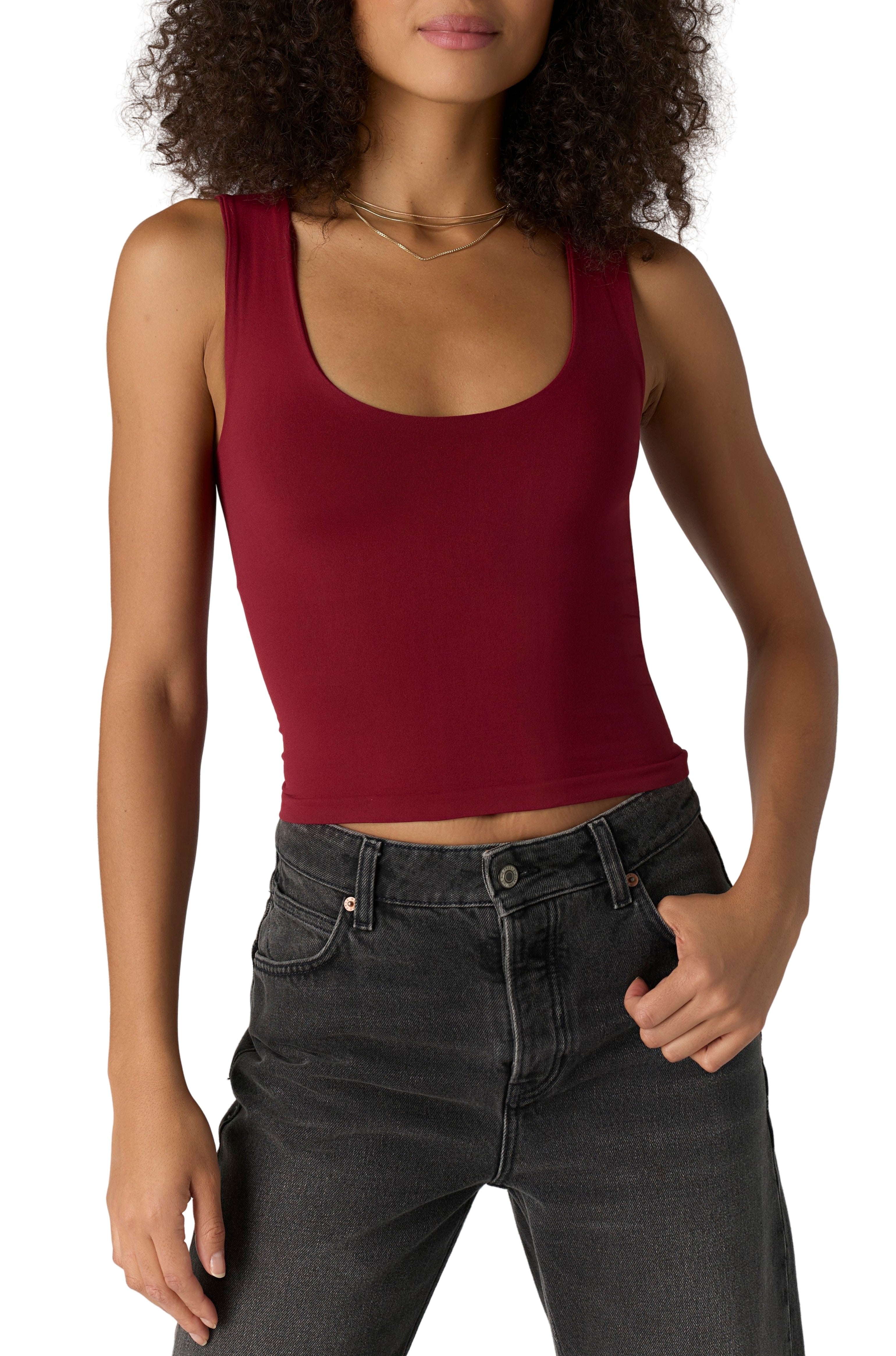 A woman wears the Free People Clean Lines Muscle Cami in pomegranate with high-waisted black jeans, one hand in her pocket, standing against a white background.