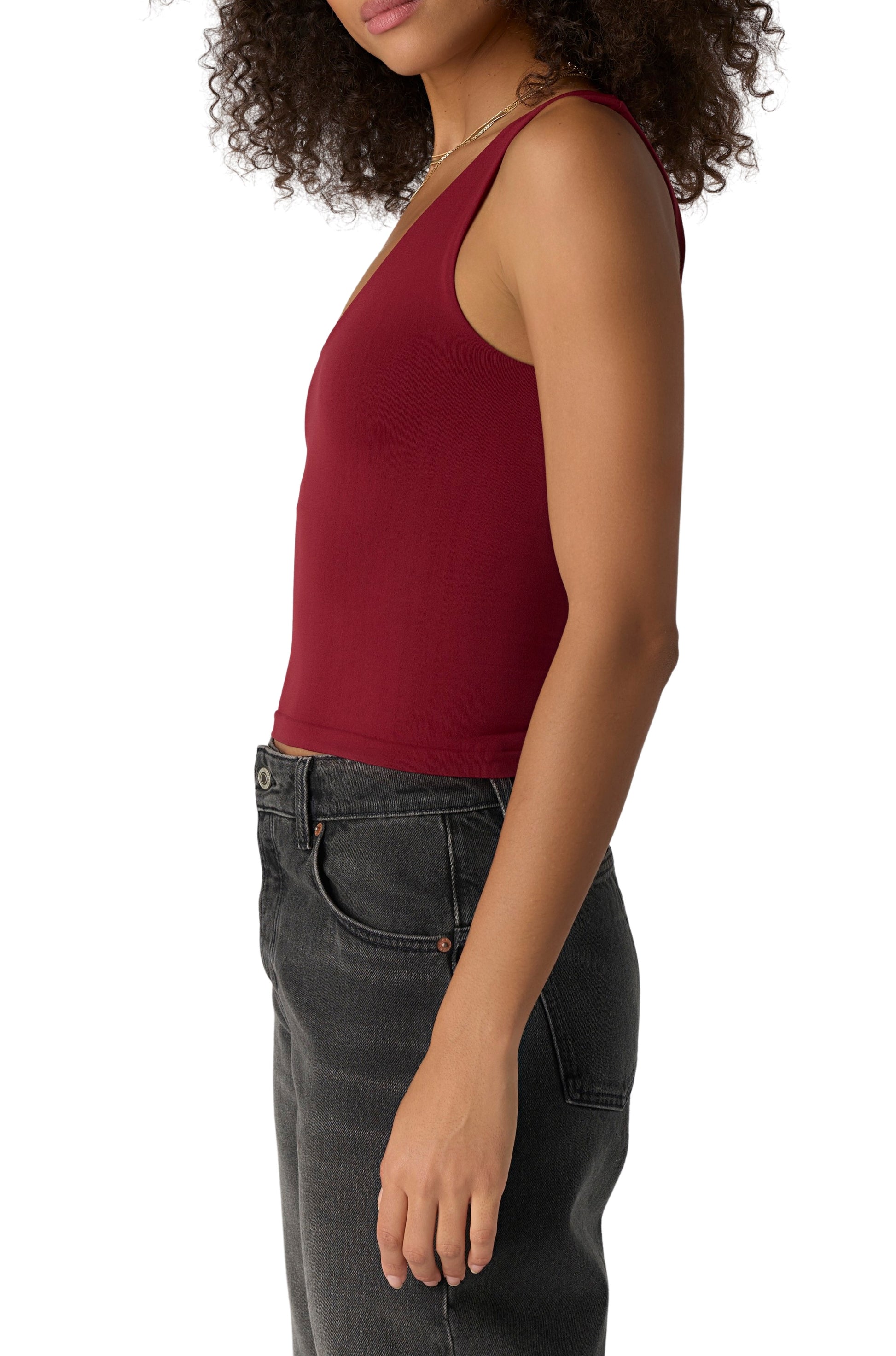 A woman with curly hair is seen from the side, wearing the Free People Clean Lines Muscle Cami in Pomegranate and high-waisted black jeans, standing against a white backdrop.