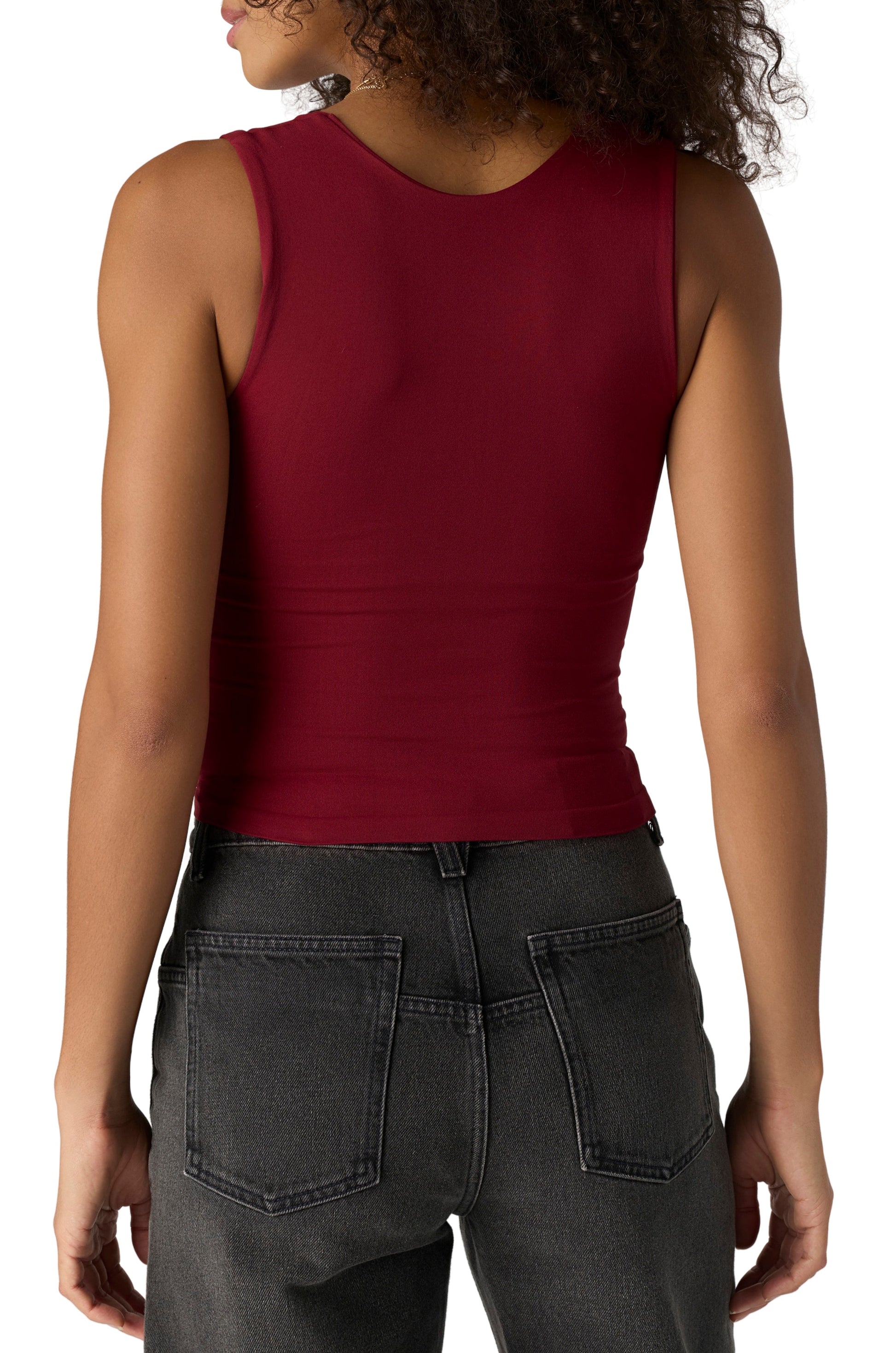 A person with curly hair is seen from the back, wearing the Free People Clean Lines Muscle Cami in Pomegranate—a sleeveless, ultra-stretchy seamless top—paired with high-waisted black jeans against a plain white background.