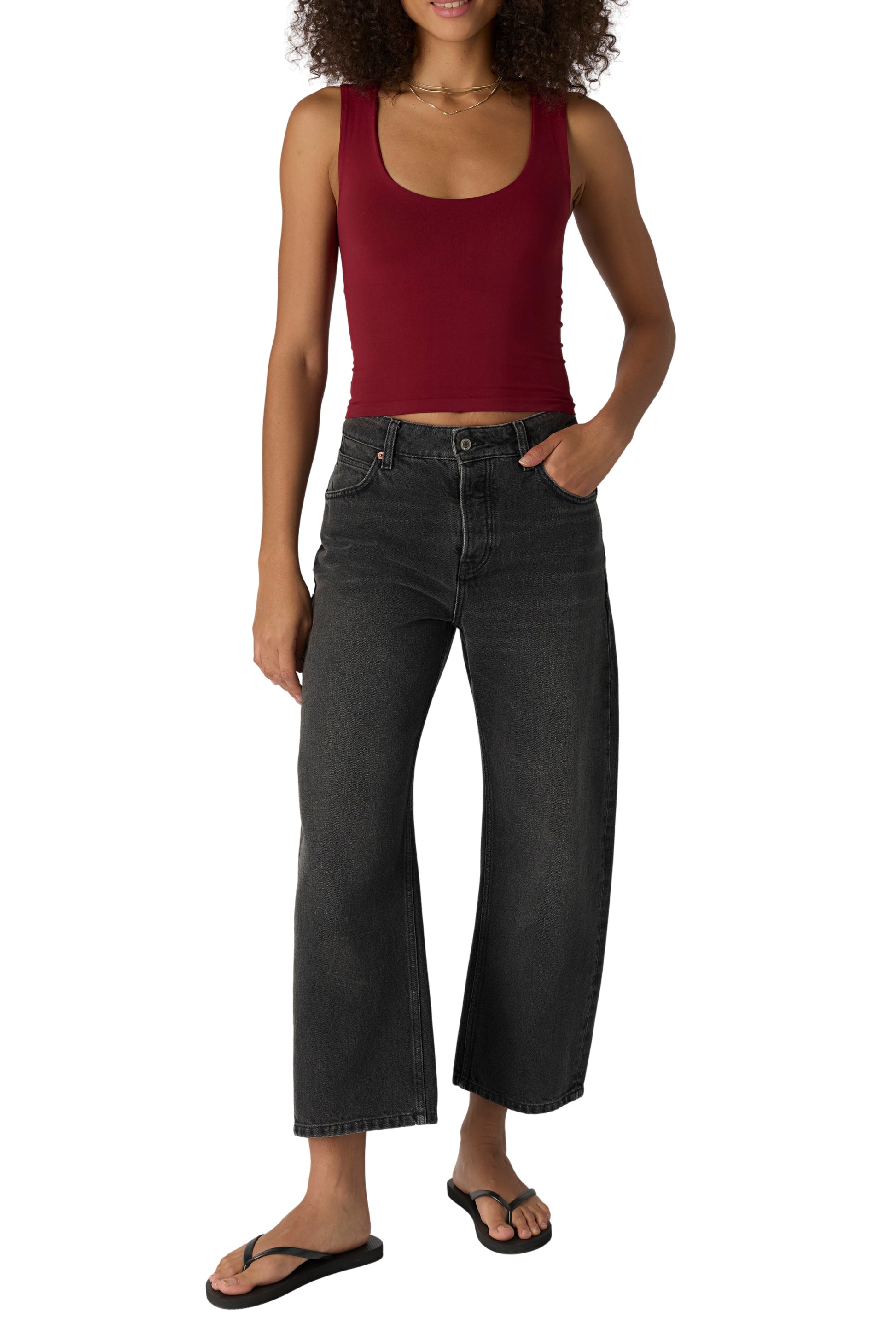 A person wearing the Free People Clean Lines Muscle Cami in Pomegranate, paired with high-waisted black wide-leg jeans and black flip-flops, stands with one hand in their pocket against a white background.