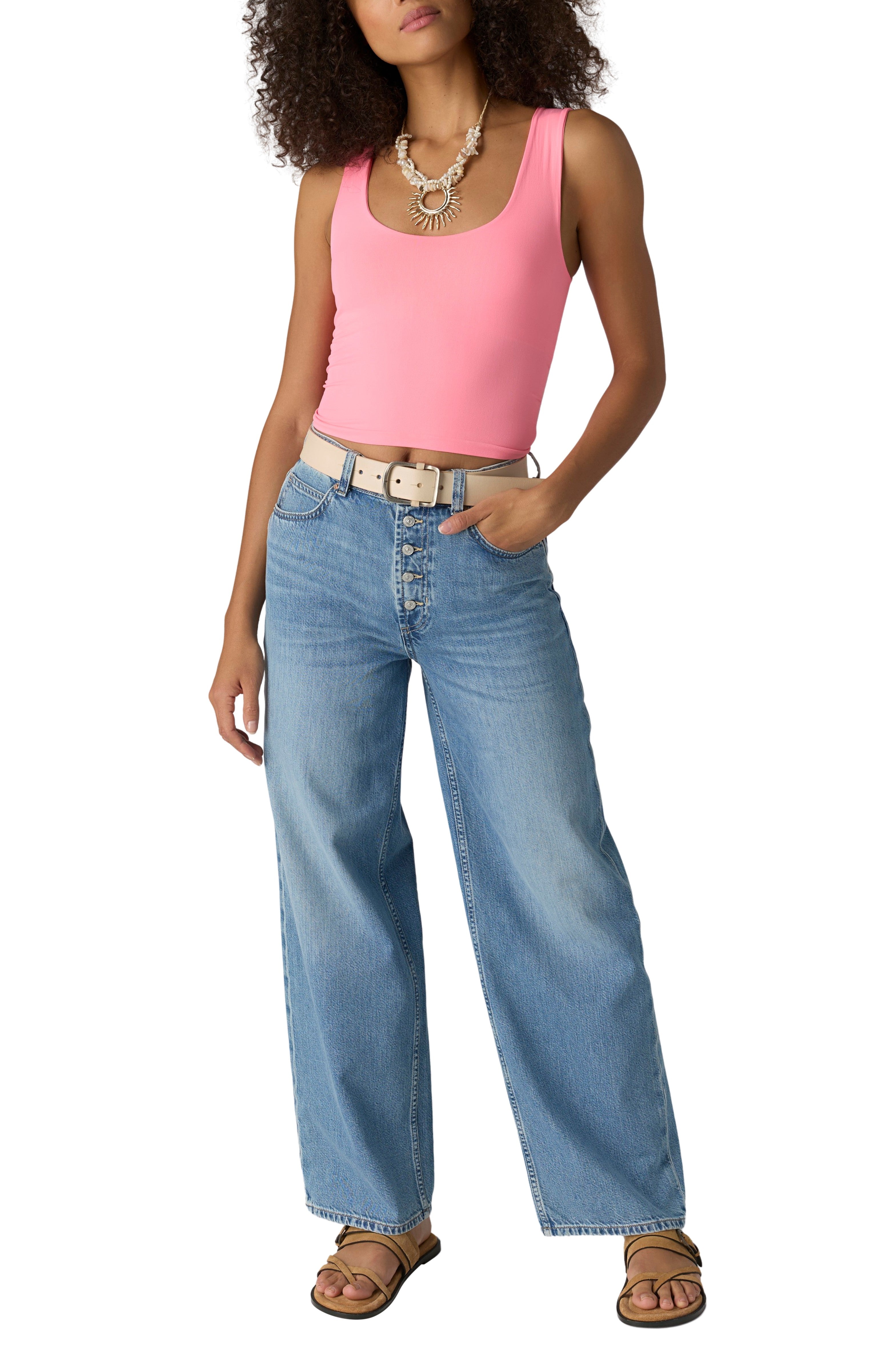 A woman wears the Free People Clean Lines Muscle Cami, sleeveless and pink, paired with light blue high-waisted wide-leg jeans, a white belt, sandals, and a statement necklace, standing with one hand in her pocket.