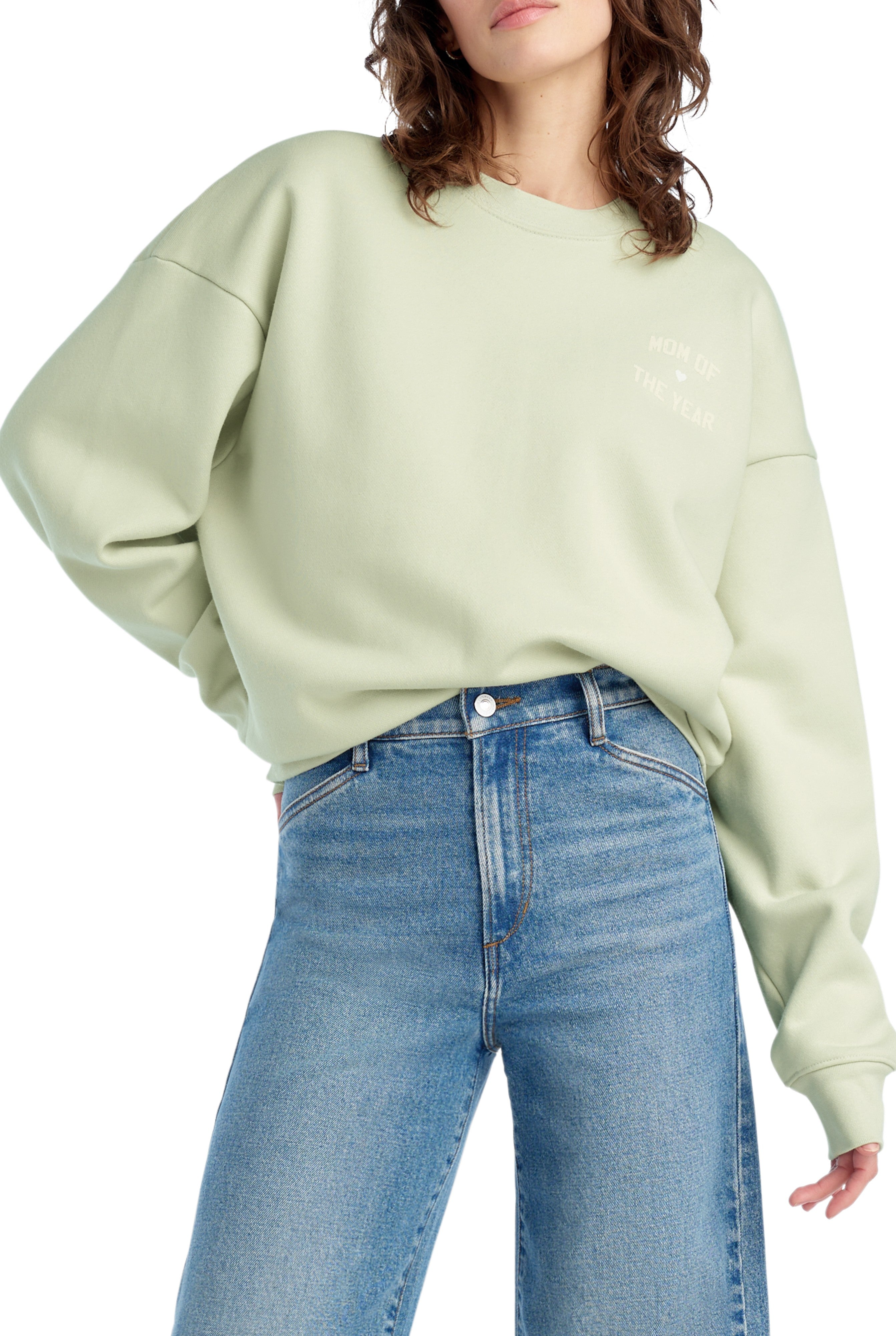 A person wears the Favorite Daughter Mom Of The Year Chest Logo sweatshirt in light green, featuring a relaxed fit and drop shoulder seams, paired with high-waisted blue jeans. The person’s face is partially out of frame.