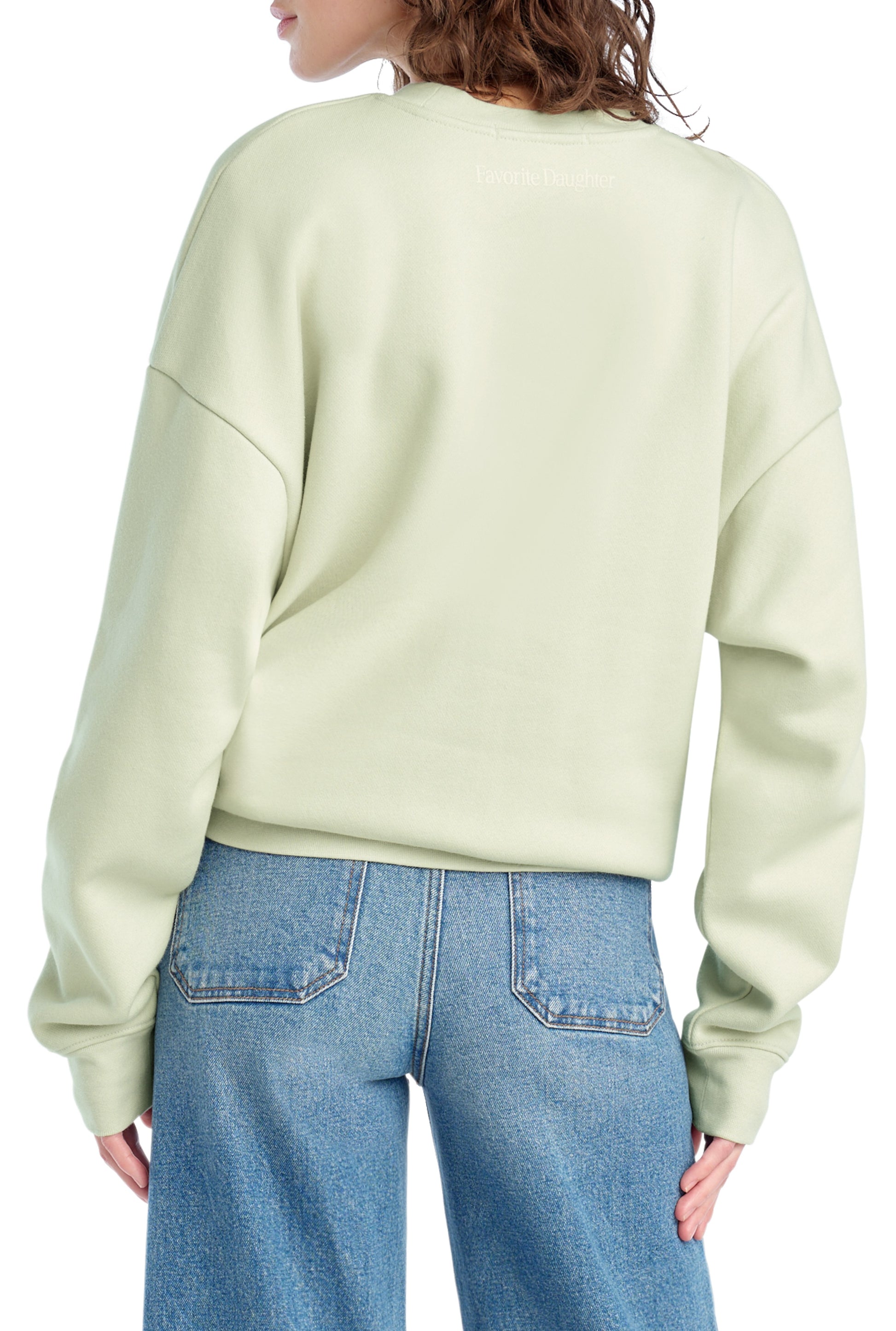 A person stands facing away, wearing the Favorite Daughter "Mom Of The Year Chest Logo" relaxed fit, light green cotton blend sweatshirt with drop shoulders and blue jeans. Only their upper back and part of their face are visible.