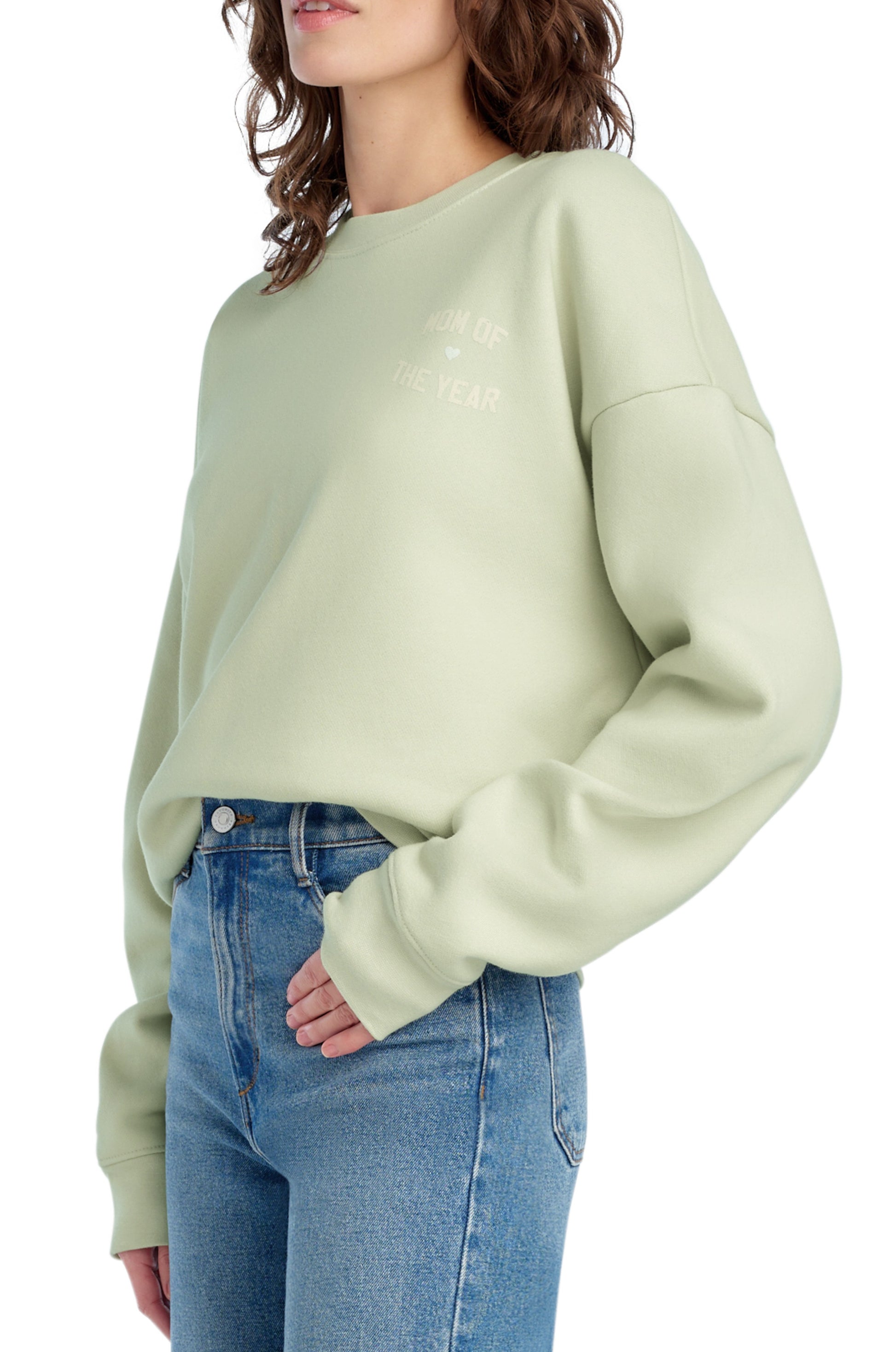 A woman with wavy brown hair wears the Favorite Daughter "Mom Of The Year Chest Logo" light green cotton blend sweatshirt and blue jeans, standing sideways with one hand in her pocket against a white background.