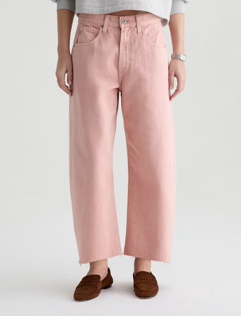 Wearing the Hattie Crop in Sulfur Sparkling Rose by AG Jeans, styled with a grey top, silver watch, and brown loafers against a plain light background.