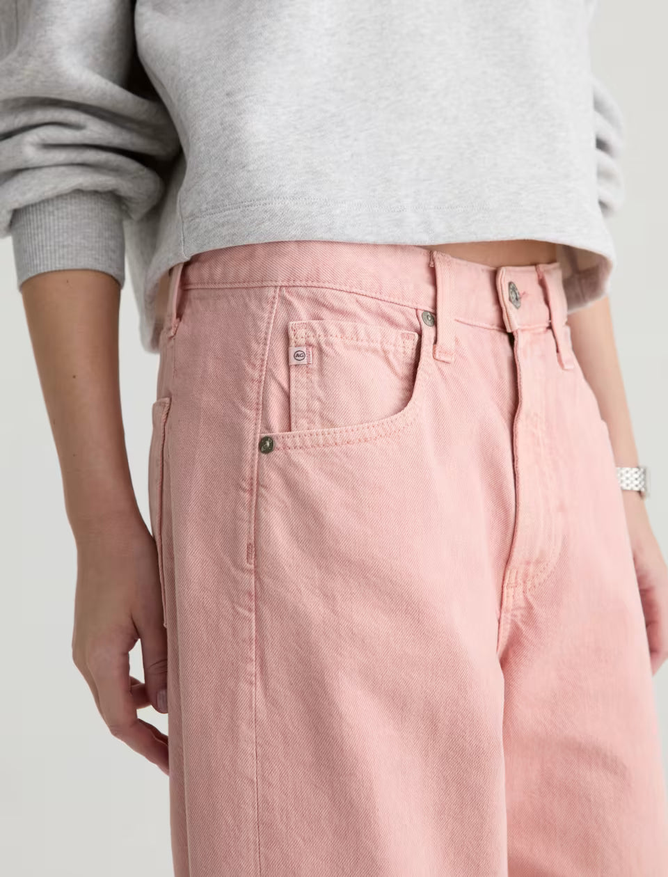 A person is shown wearing AG Jeans' Hattie Crop In Sulfur Sparkling Rose—high-waisted, loose-fitting pink jeans—paired with a cropped gray sweatshirt. The image displays the torso and upper legs, with one hand at their side.