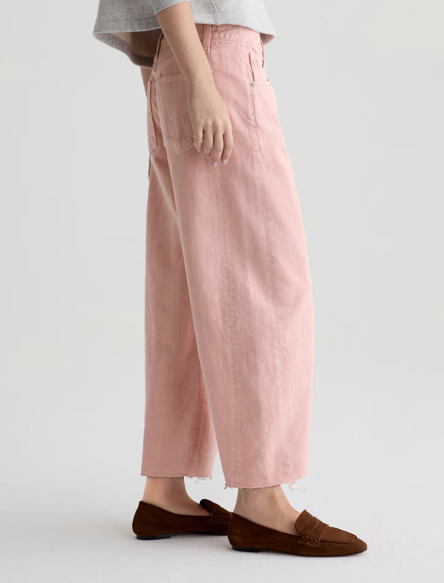 A person wearing the Hattie Crop In Sulfur Sparkling Rose by AG Jeans with brown loafers stands sideways against a plain light background, showing only the lower half of their body.