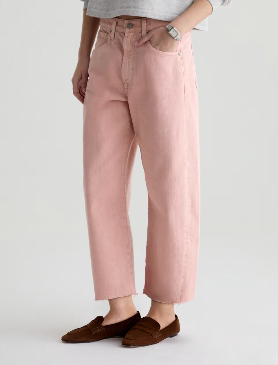 A person wearing AG Jeans' Hattie Crop in Sulfur Sparkling Rose, a light grey top, silver wristwatch, and brown loafers stands against a plain light background with hands in pockets.
