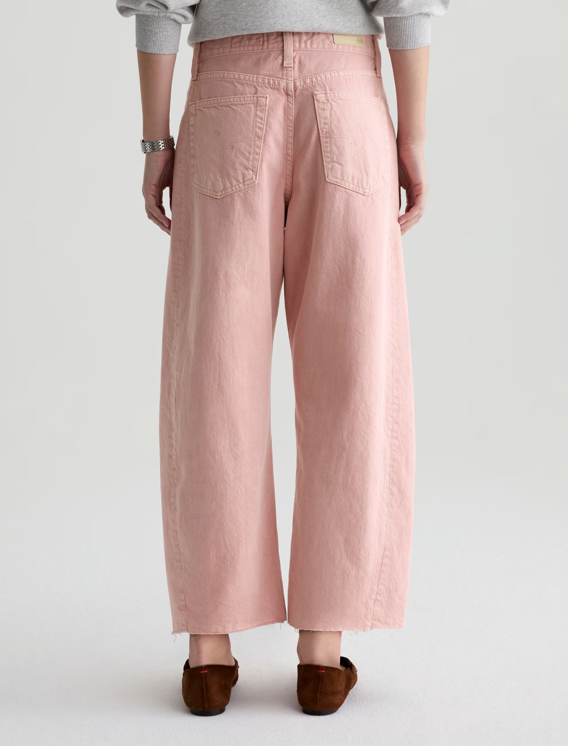 A person is seen from the back wearing AG Jeans' Hattie Crop in Sulfur Sparkling Rose—wide-leg, light pink jeans with frayed hems—paired with a light gray top, brown flats, and a silver bracelet against a plain light background.