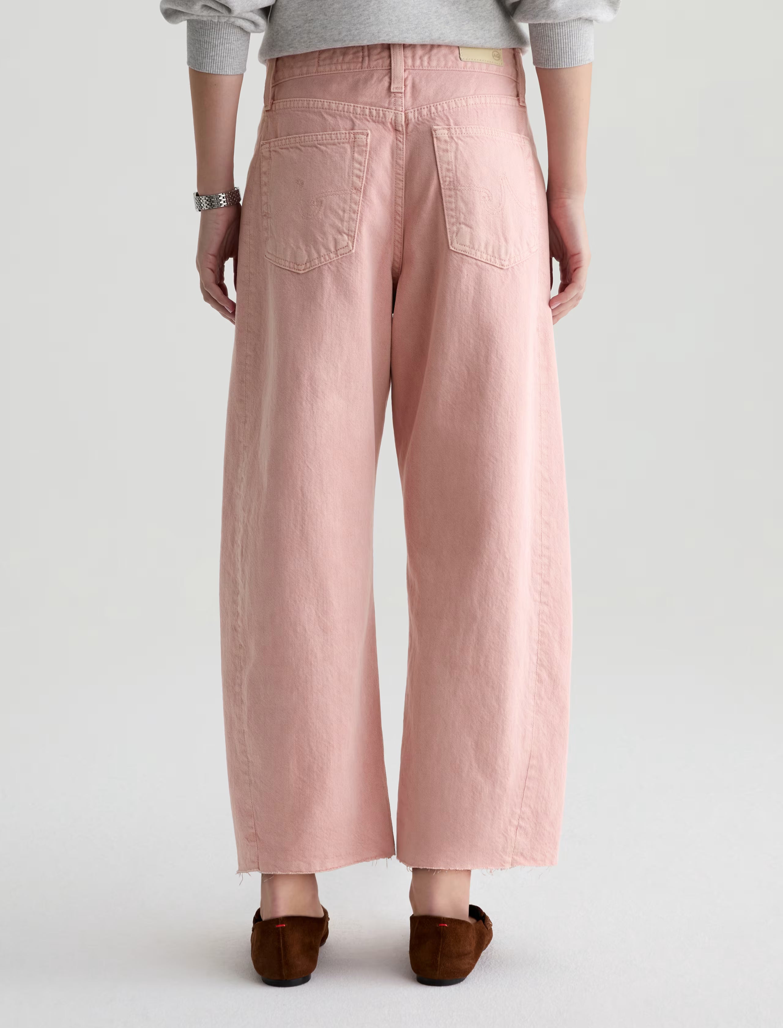 A person is seen from the back wearing AG Jeans' Hattie Crop in Sulfur Sparkling Rose—wide-leg, light pink jeans with frayed hems—paired with a light gray top, brown flats, and a silver bracelet against a plain light background.
