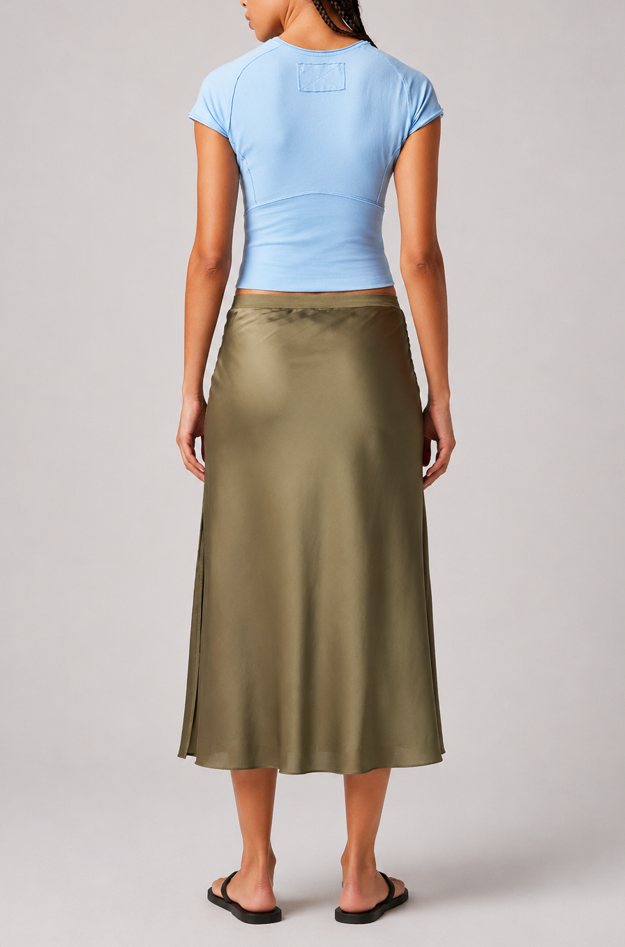 A woman faces away, wearing the Protagonist Tee by Free People in light blue, paired with an olive green satin midi skirt and black sandals, set against a plain light background.