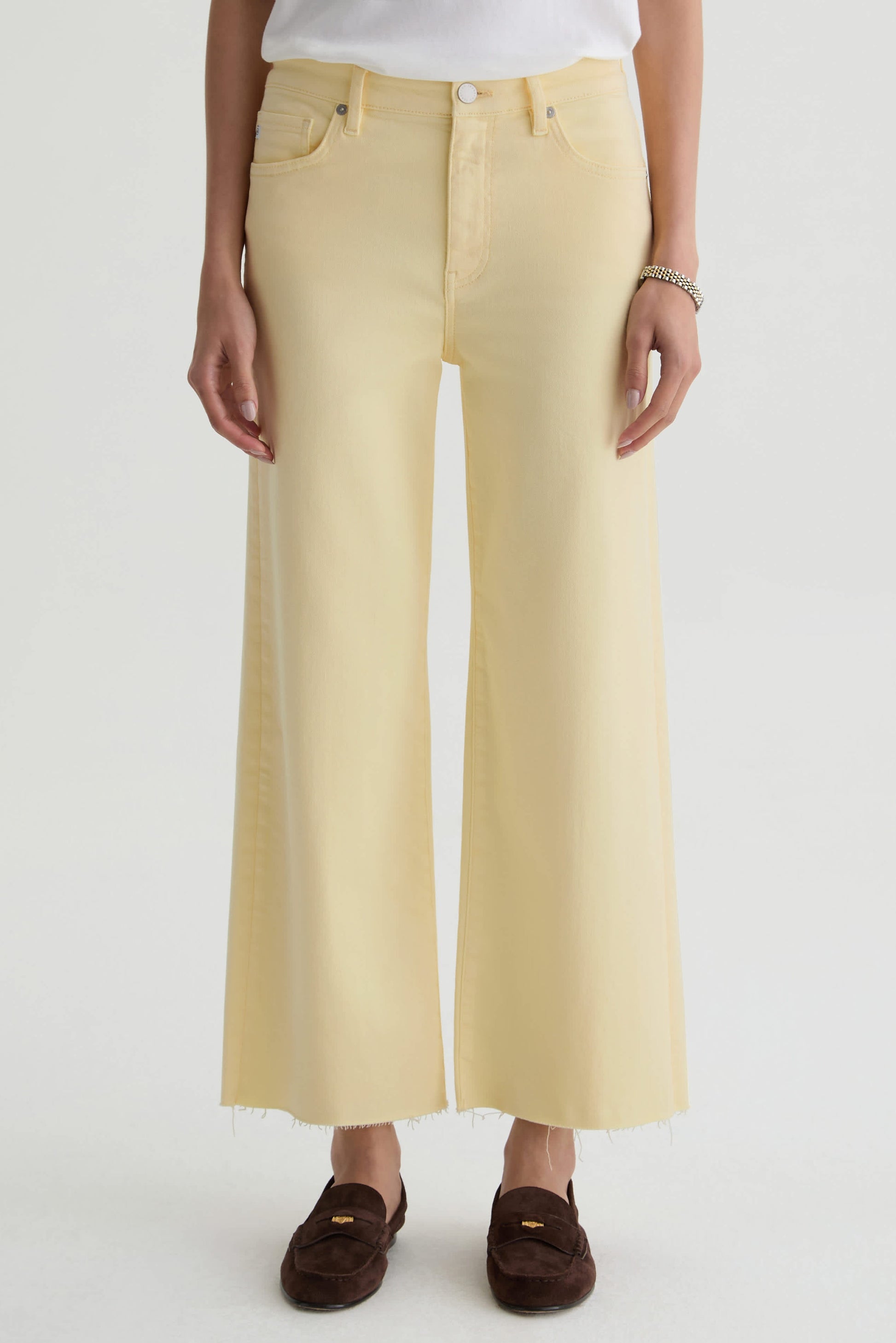 Wearing the AG Jeans Saige Wide Leg Crop in sunlight yellow cloud soft denim with raw hems, a white top, bracelet, and brown loafers, a person stands against a plain light background.
