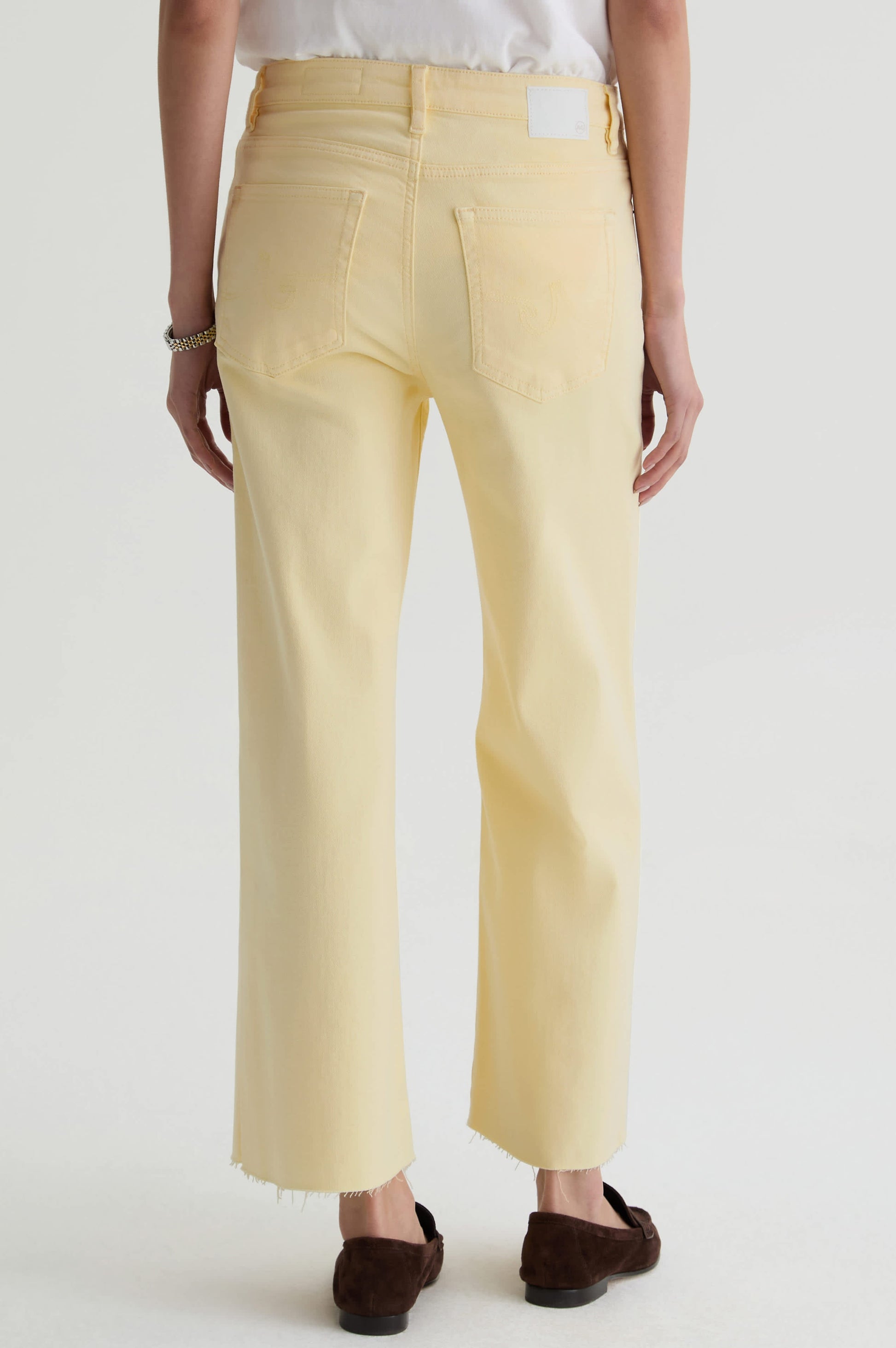 A person stands facing away, wearing AG Jeans' Saige Wide Leg Crop in sunlight yellow, made from cloud soft denim with frayed hems. They pair it with a white top and brown shoes, set against a plain white background.