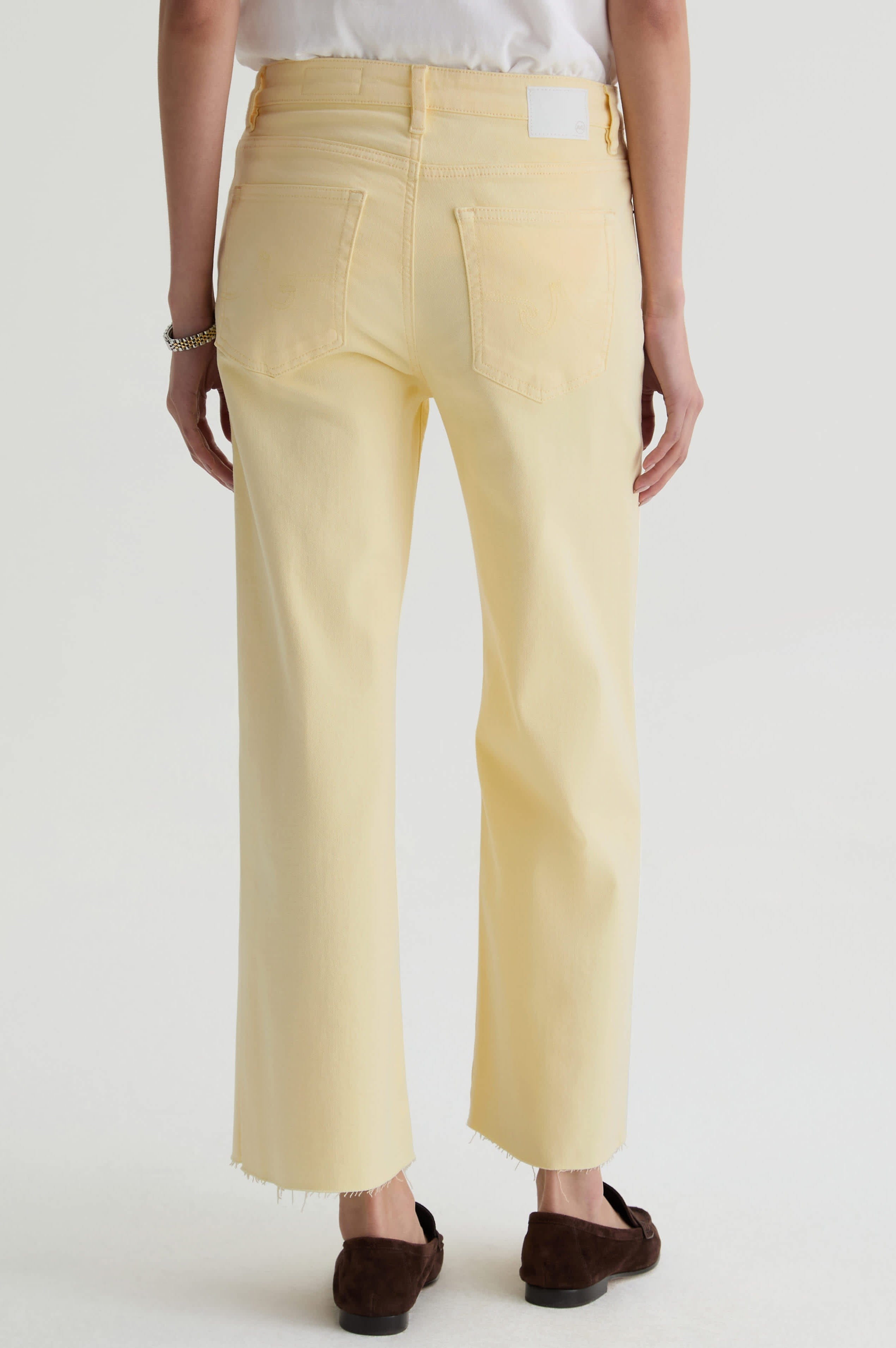 A person stands facing away, wearing AG Jeans' Saige Wide Leg Crop in sunlight yellow, made from cloud soft denim with frayed hems. They pair it with a white top and brown shoes, set against a plain white background.