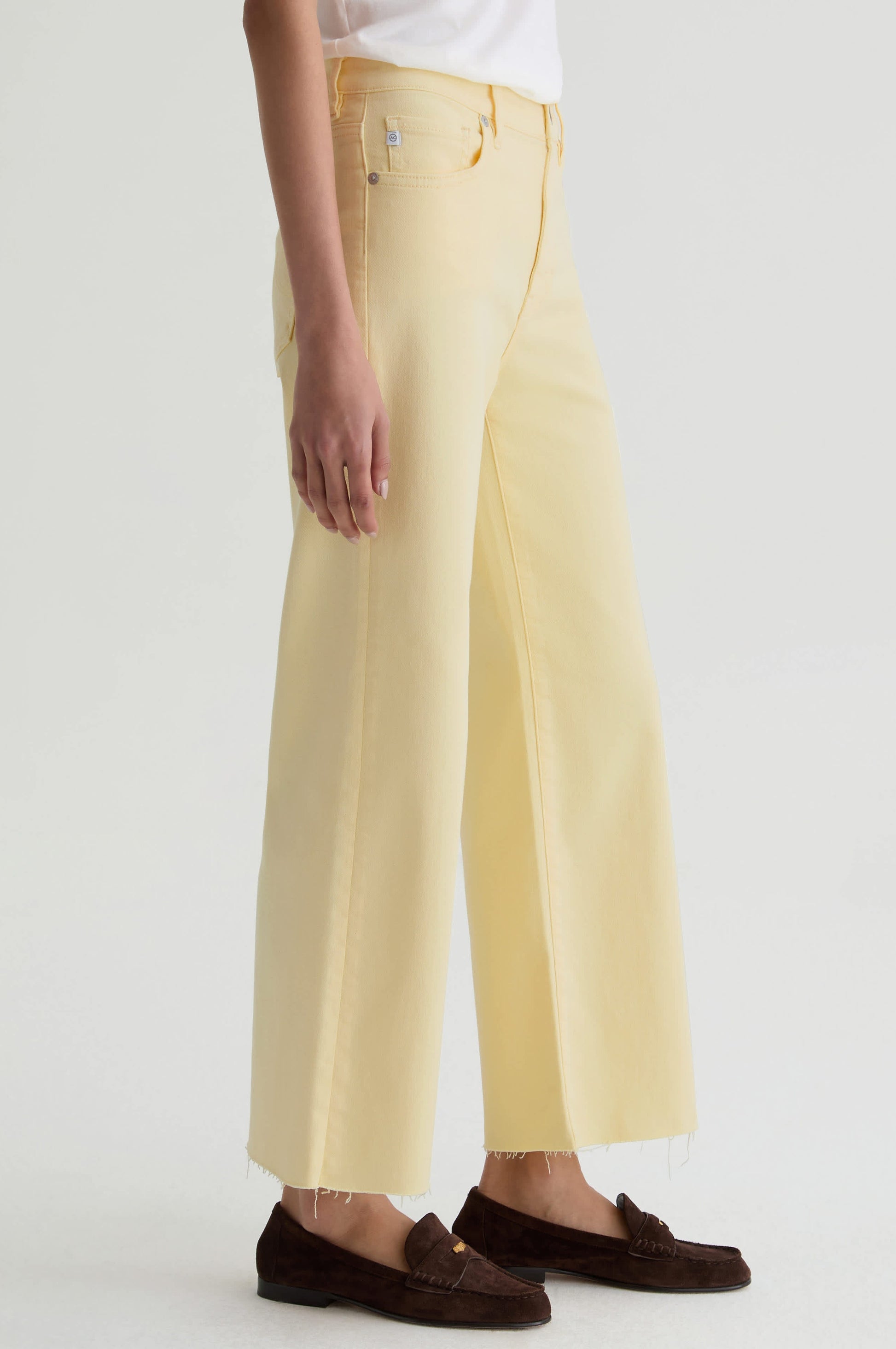 Someone wearing the Saige Wide Leg Crop jeans by AG Jeans in sunlight yellow cloud soft denim, paired with a white top and brown loafers, stands against a plain white background.