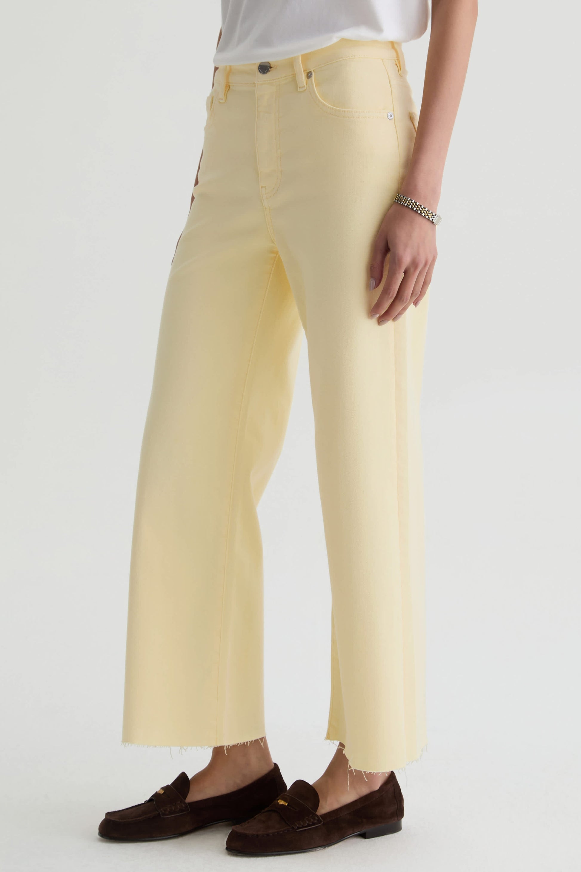 Wearing AG Jeans’ Saige Wide Leg Crop in sunlight yellow cloud soft denim with raw hems, a white top, silver bracelet, and brown loafers, a person stands against a plain white background.