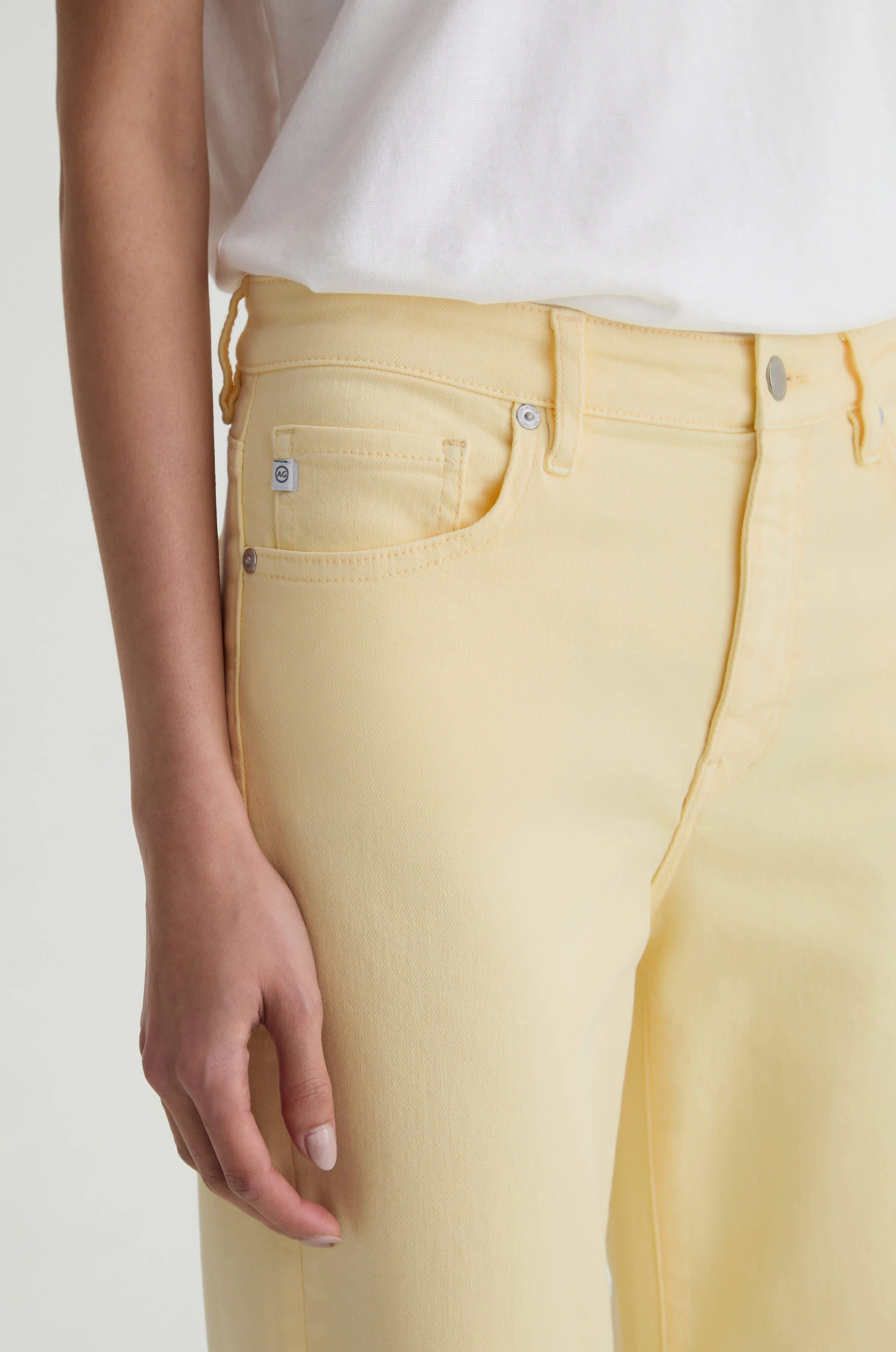 A person wears Saige Wide Leg Crop jeans by AG Jeans in sunlight yellow cloud soft denim and a white t-shirt, shown waist to mid-thigh with one arm relaxed at their side.