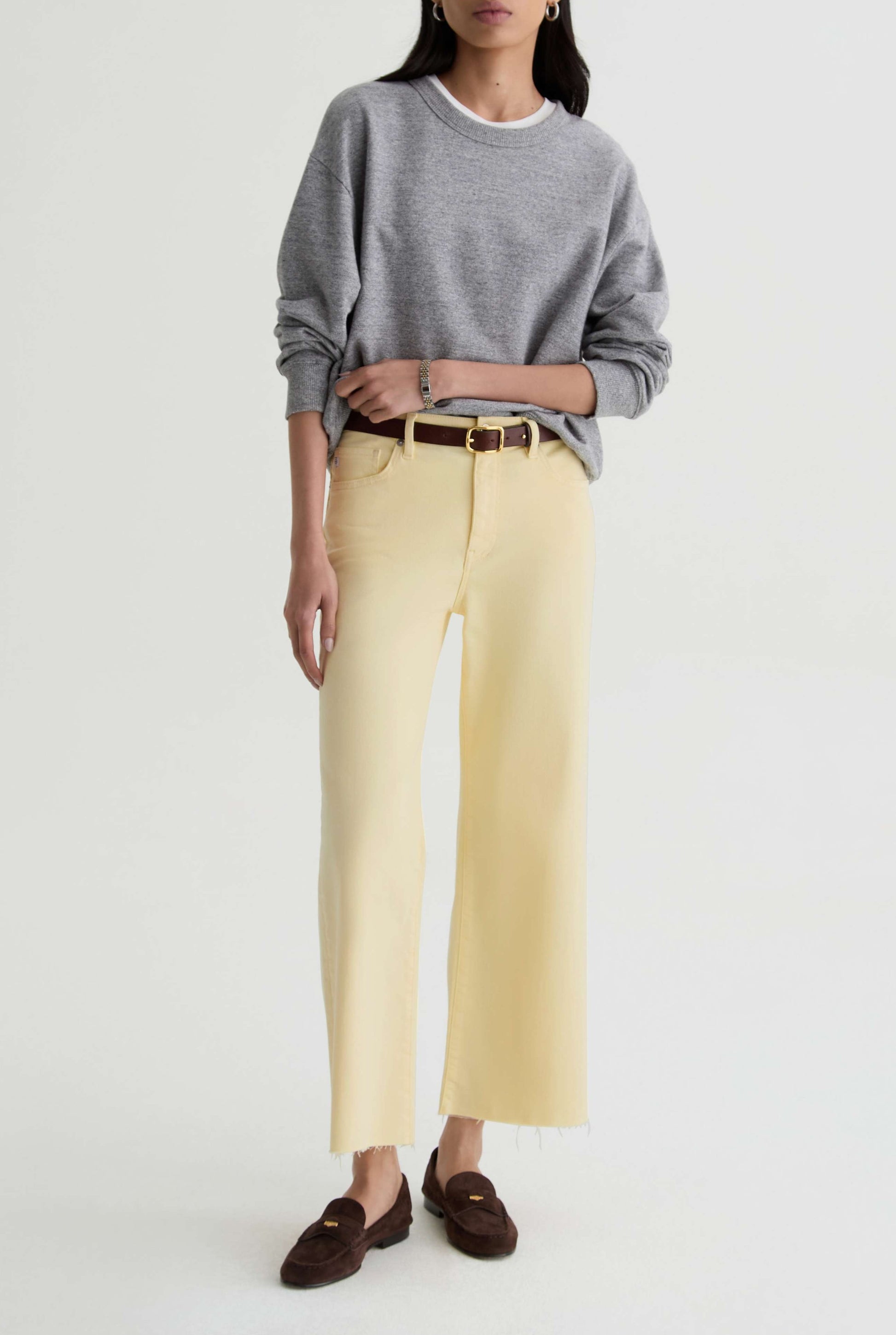 A woman stands against a plain background wearing a gray long-sleeve top, AG Jeans’ Saige Wide Leg Crop in sunlight yellow cloud soft denim, a brown belt, and dark brown loafers. Her left hand rests on her waist; her right arm is relaxed.