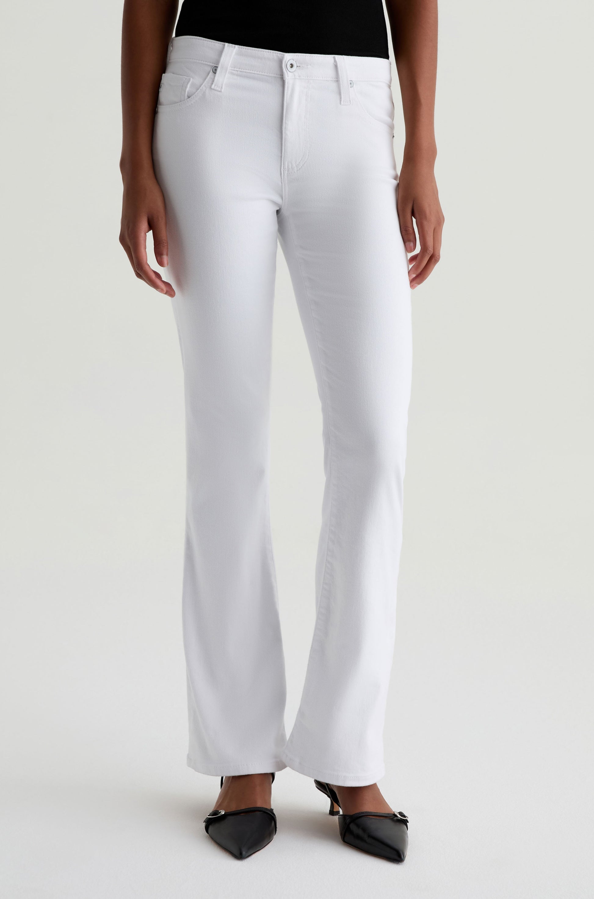A person wears the AG Jeans Angel white low-rise bootcut jeans with black pointed-toe shoes, standing against a plain light background. Their relaxed arms are at their sides and only part of their upper body is visible.